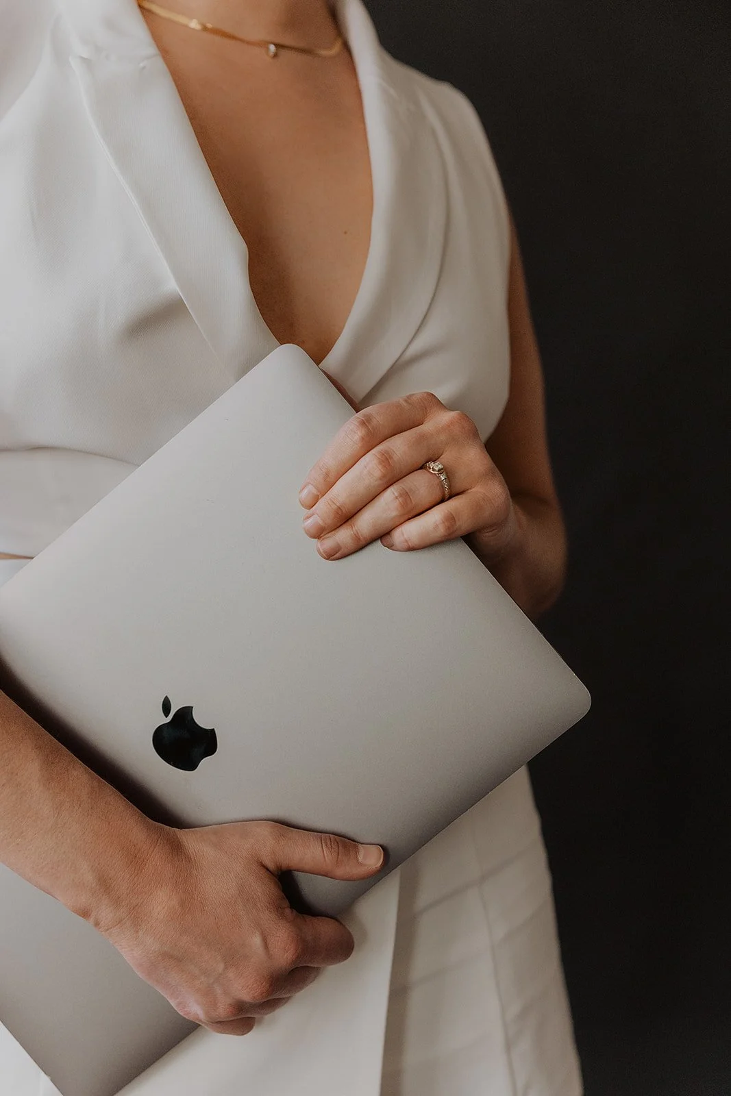 A woman in a white sleeveless blouse holding a closed silver MacBook laptop with both hands, one hand on the top and the other on the side, wearing a wedding ring and a thin necklace with a small pendant.