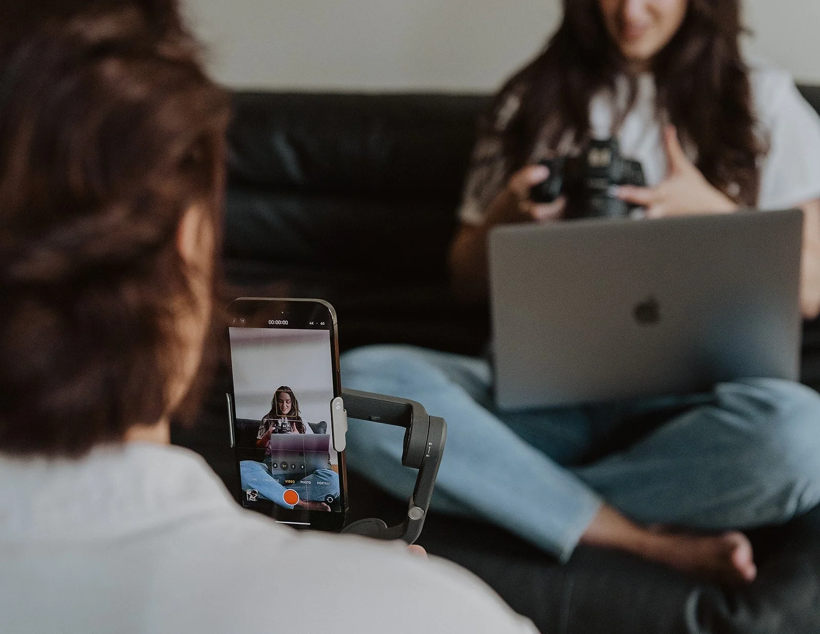 Person recording a woman sitting on a couch, who is holding a camera and smiling