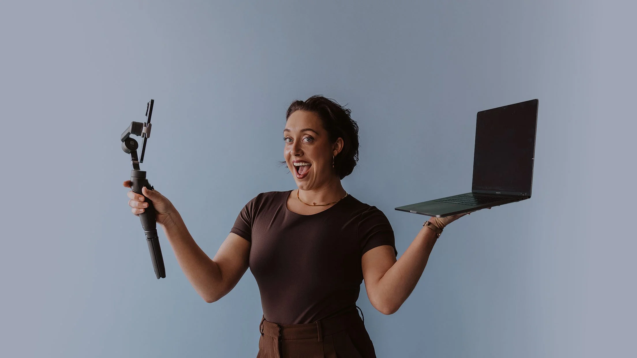 Woman holding a selfie stick in one hand and a laptop in the other, smiling and looking excited against a plain background.