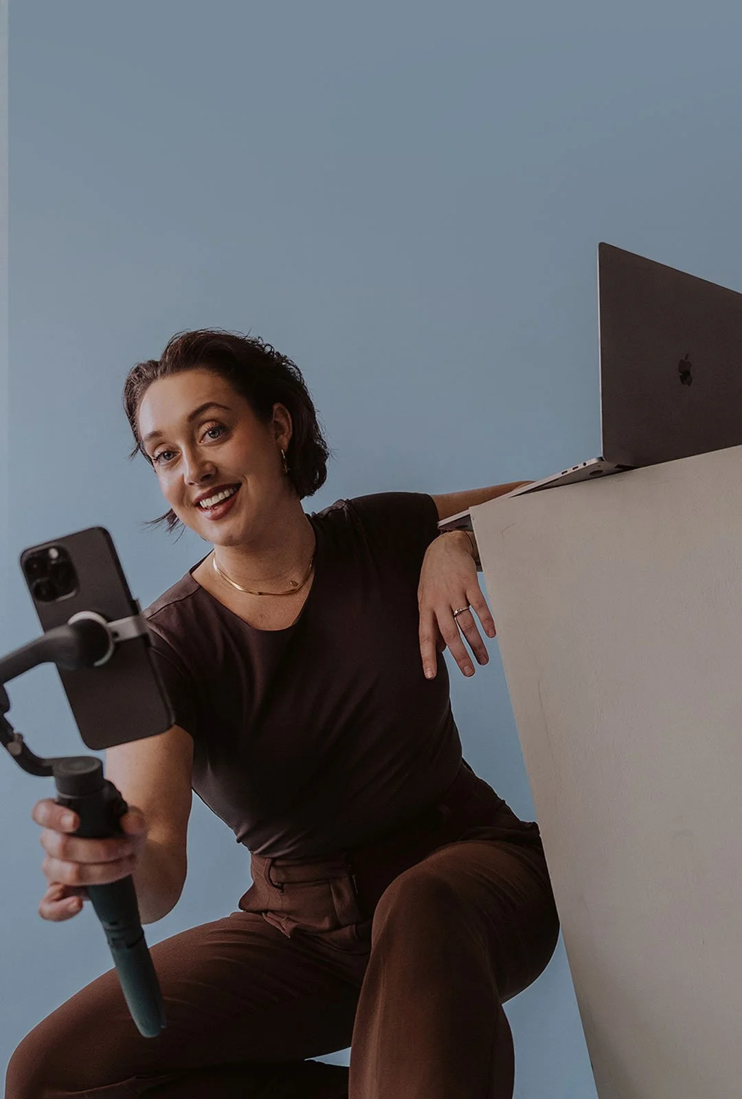 Woman smiling and recording a video with a smartphone on a stabilizer, with a laptop on a table in front of her, against a light blue background.