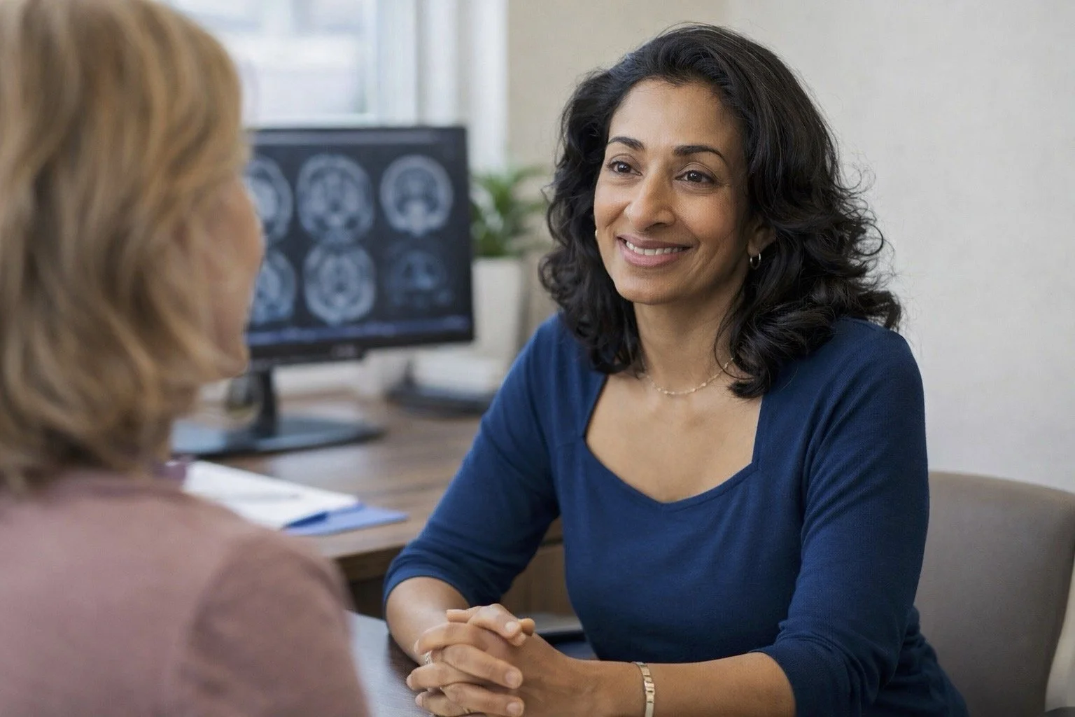 A woman with dark curly hair smiling and talking to another woman with blonde hair in an office setting with brain scans on a computer monitor in the background.
