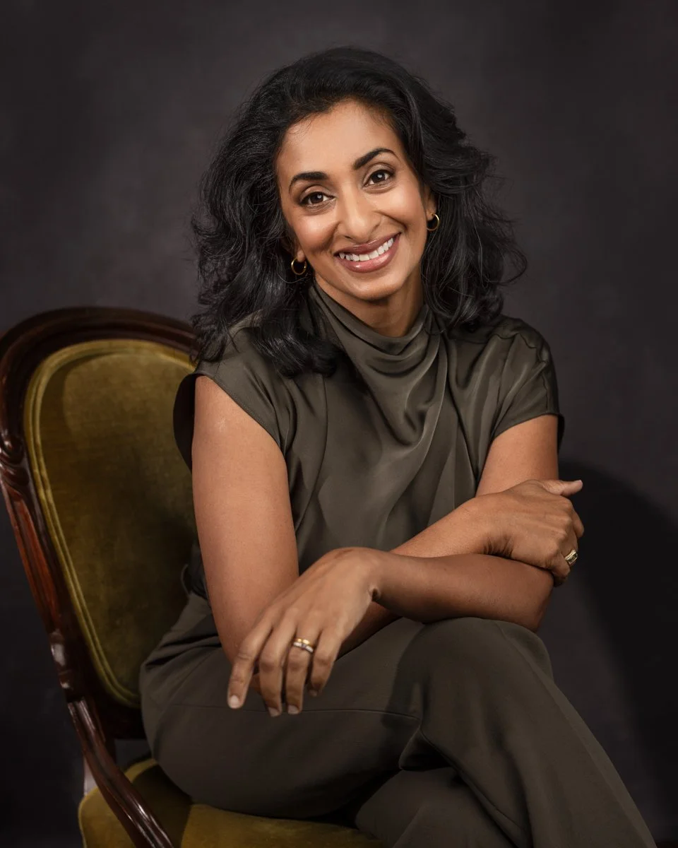 A woman with black wavy hair smiling, seated on a vintage green and wood chair against a dark background.