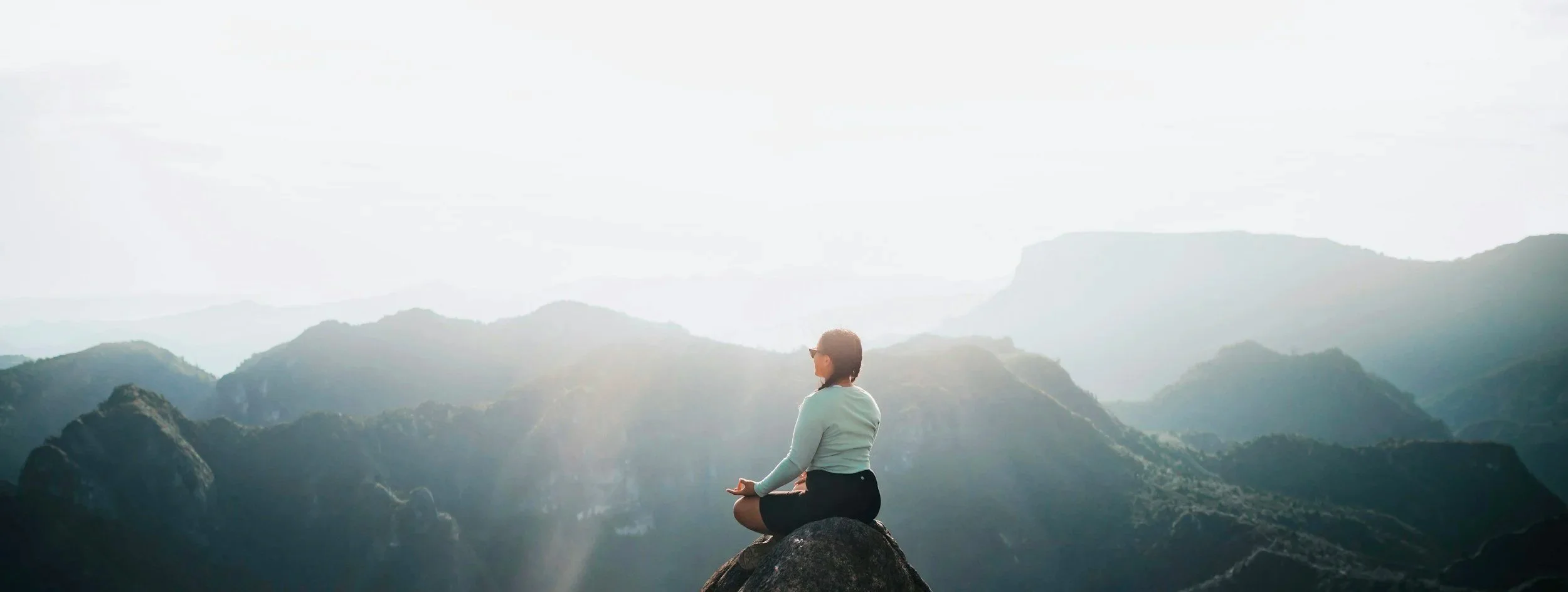 Woman practicing yoga and meditating on a large rock overlooking a mountain landscape at sunrise or sunset.