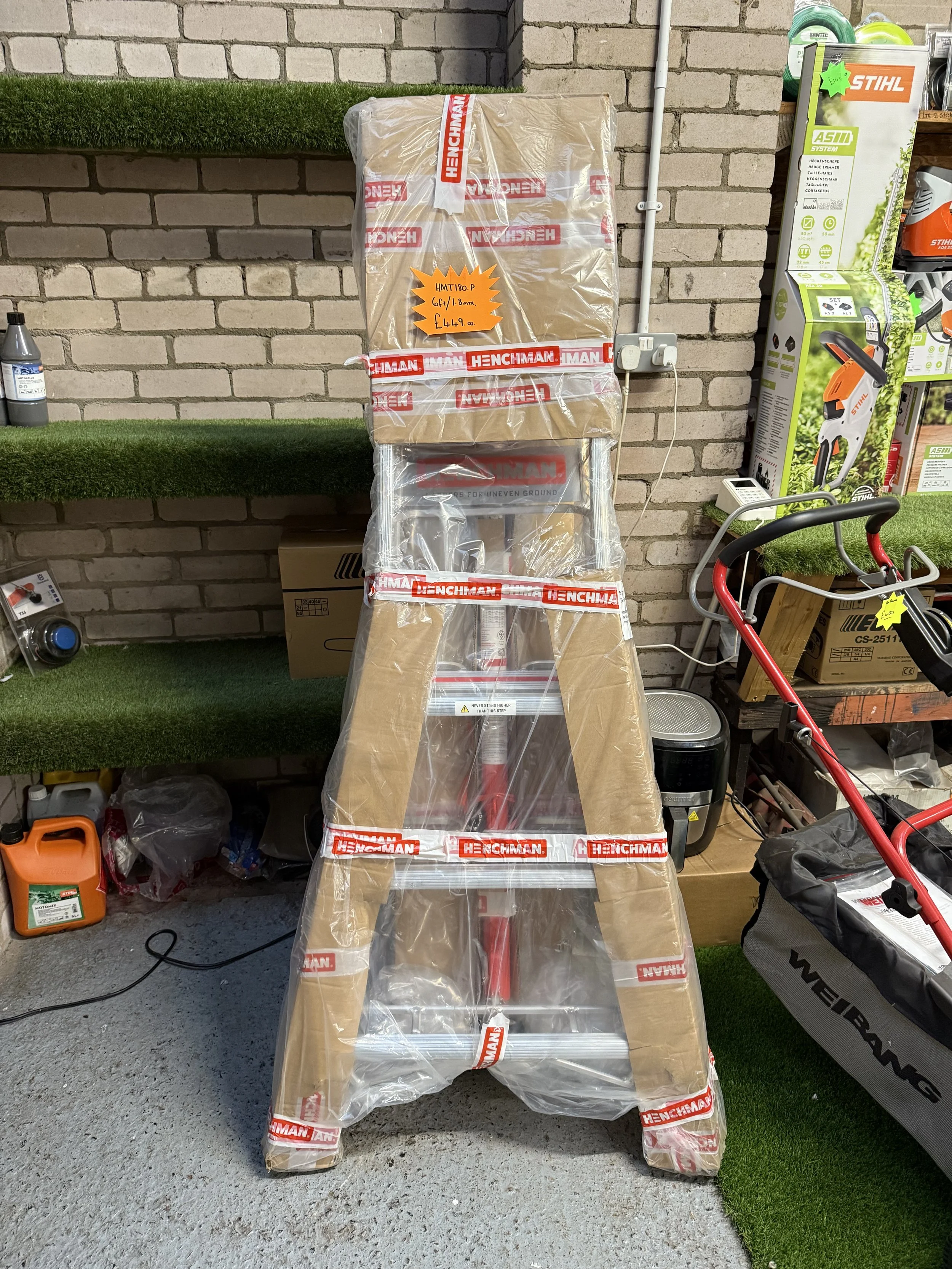 A ladder wrapped in plastic and cardboard in a hardware store. The ladder has red and silver parts. It is placed on a concrete floor near shelves with various tools and products, against a brick wall.