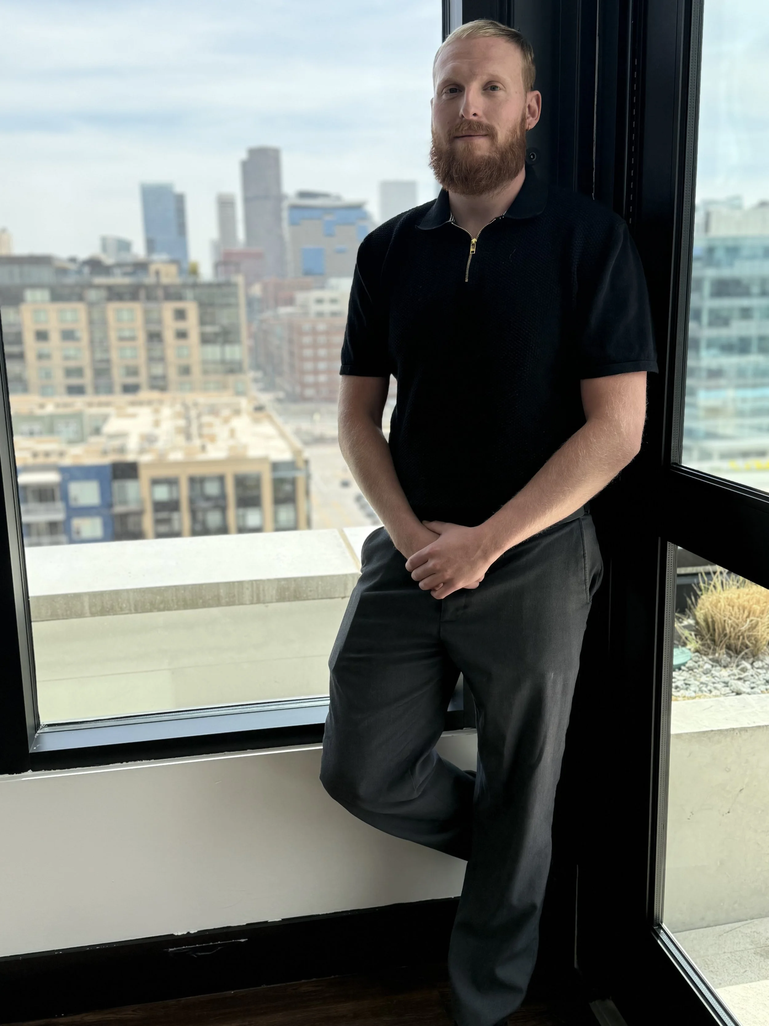 A man with a beard and light hair standing near a large window overlooking a city skyline, wearing a black polo shirt and dark pants.