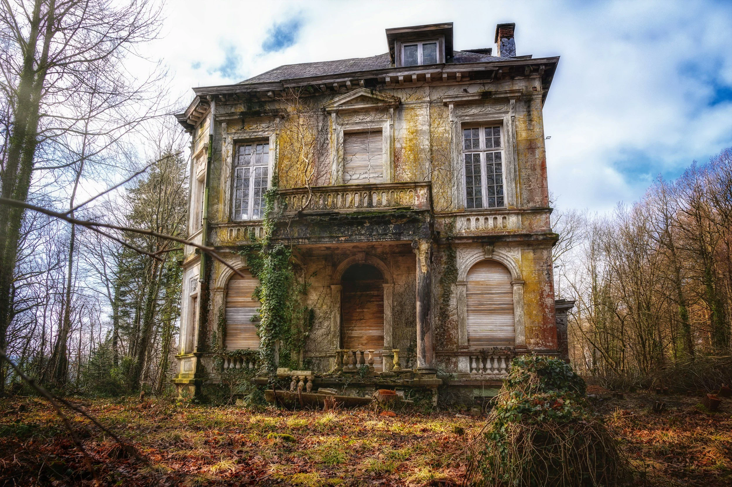 An abandoned, dilapidated mansion with broken windows, overgrown vines, and a neglected yard surrounded by leafless trees under a cloudy sky.