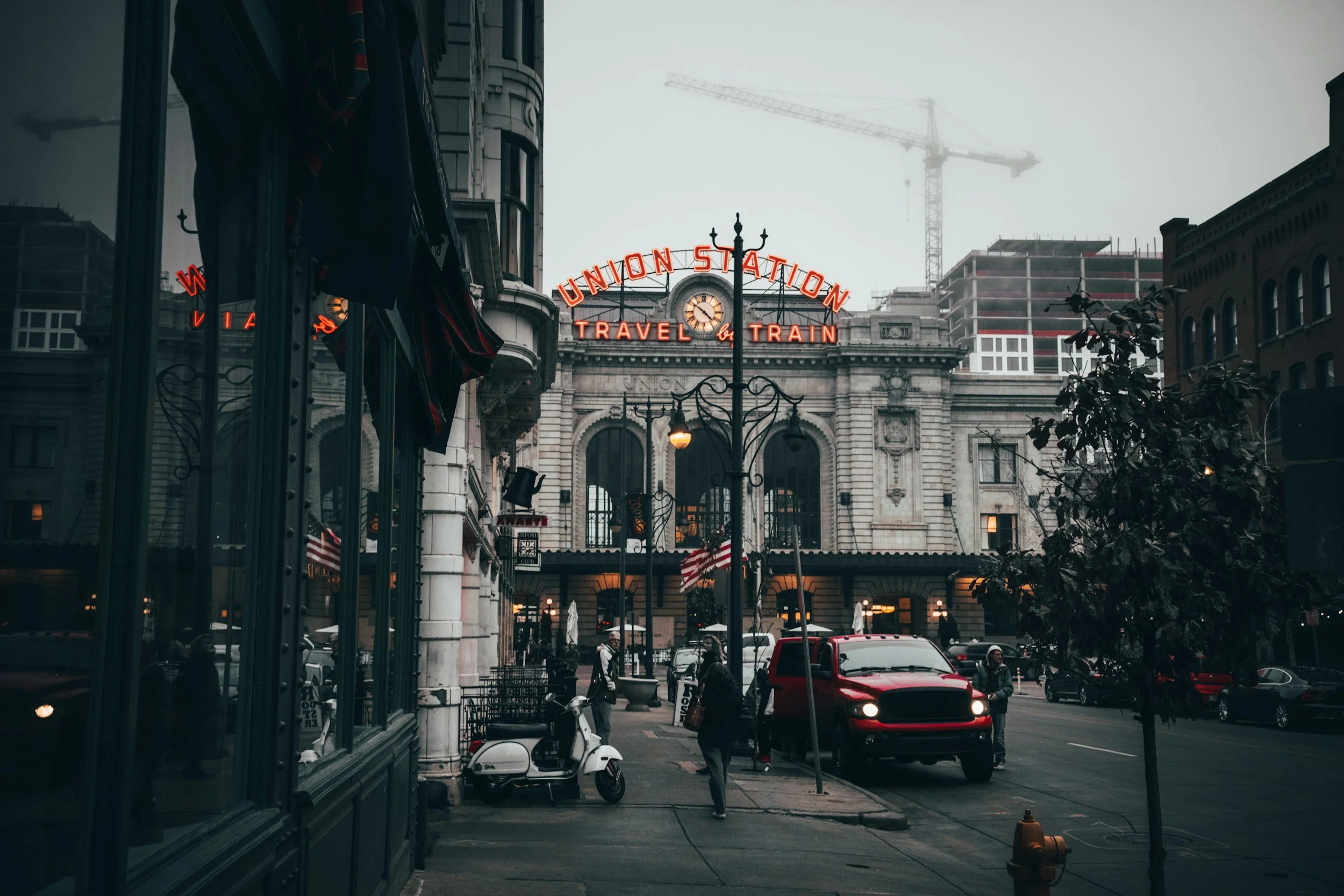 Historic train station with a large sign reading 'Union Station, Travel by Train' in red neon lights, amidst city buildings and construction crane in the background. Street scene with pedestrians, a red truck, and American flags.