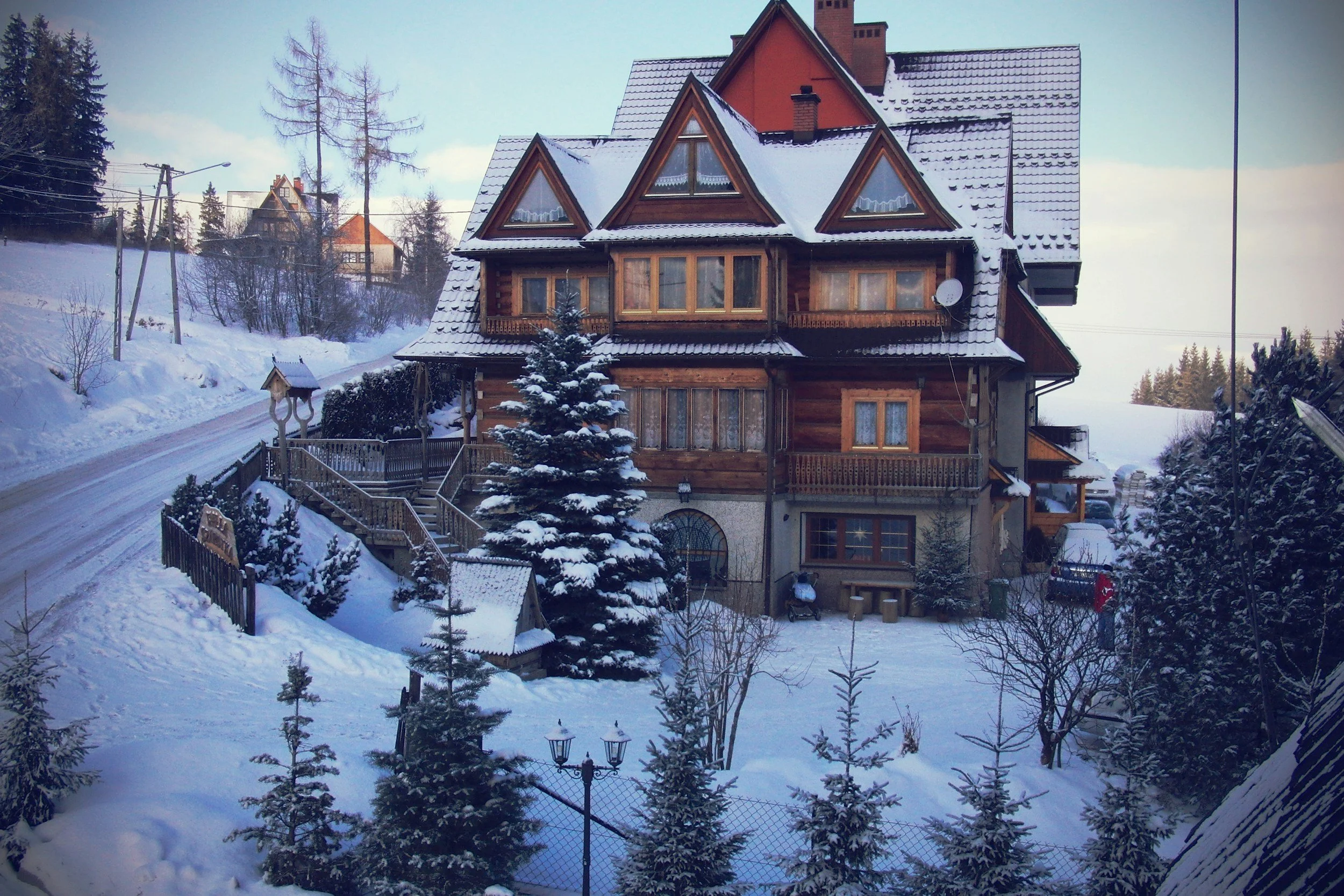 A multi-story wooden house with a snowy roof, surrounded by snow-covered trees and a fence. The scene is set in a winter landscape with snow on the ground and on the rooftops.