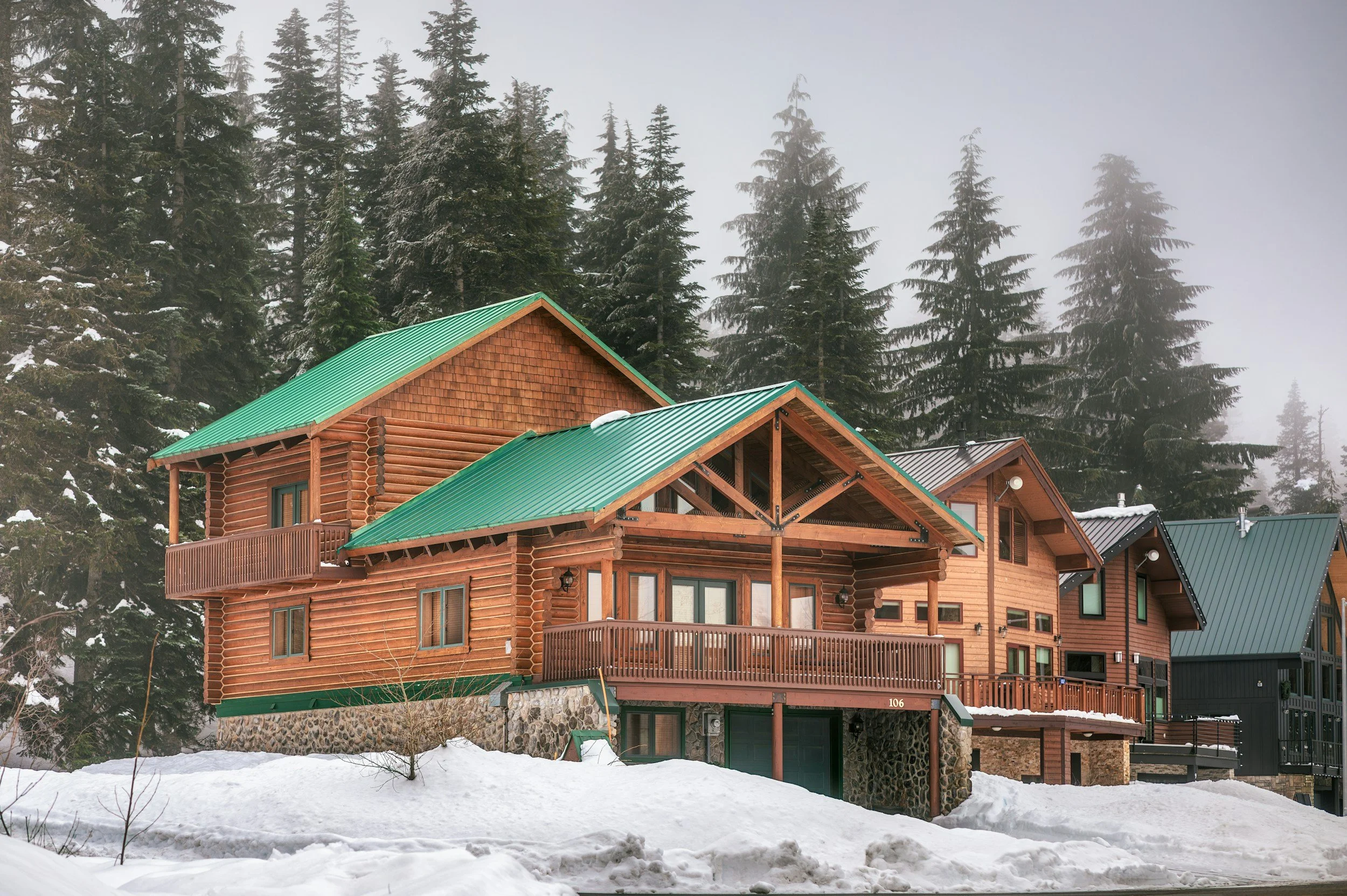 A wooden mountain house with a green metal roof, surrounded by snow and tall pine trees in the background.