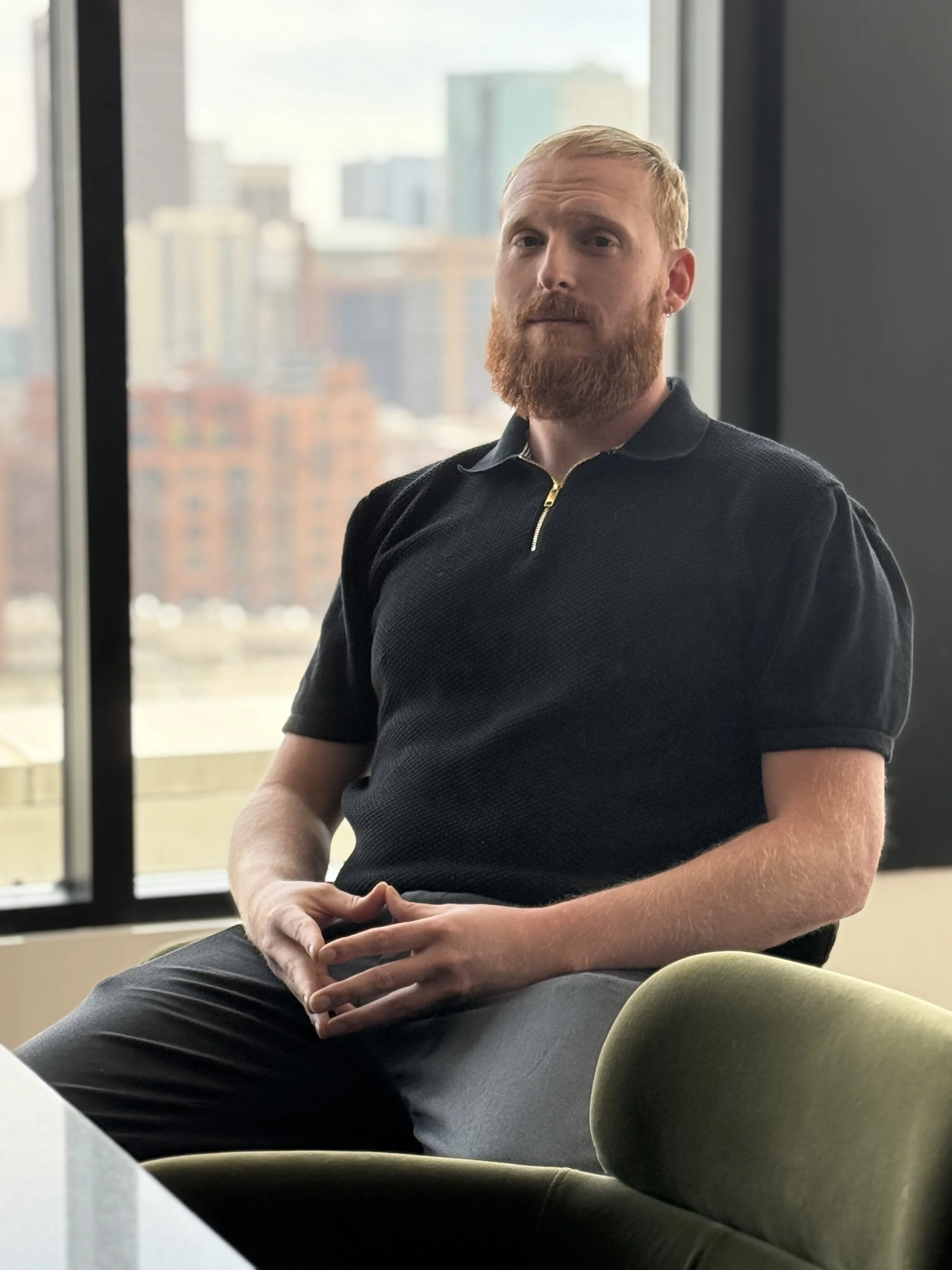 A man with a red beard and short hair sitting in a modern office, with large windows showing a cityscape in the background. He is wearing a black short-sleeved polo shirt with a zipper collar and has his hands resting on his lap.