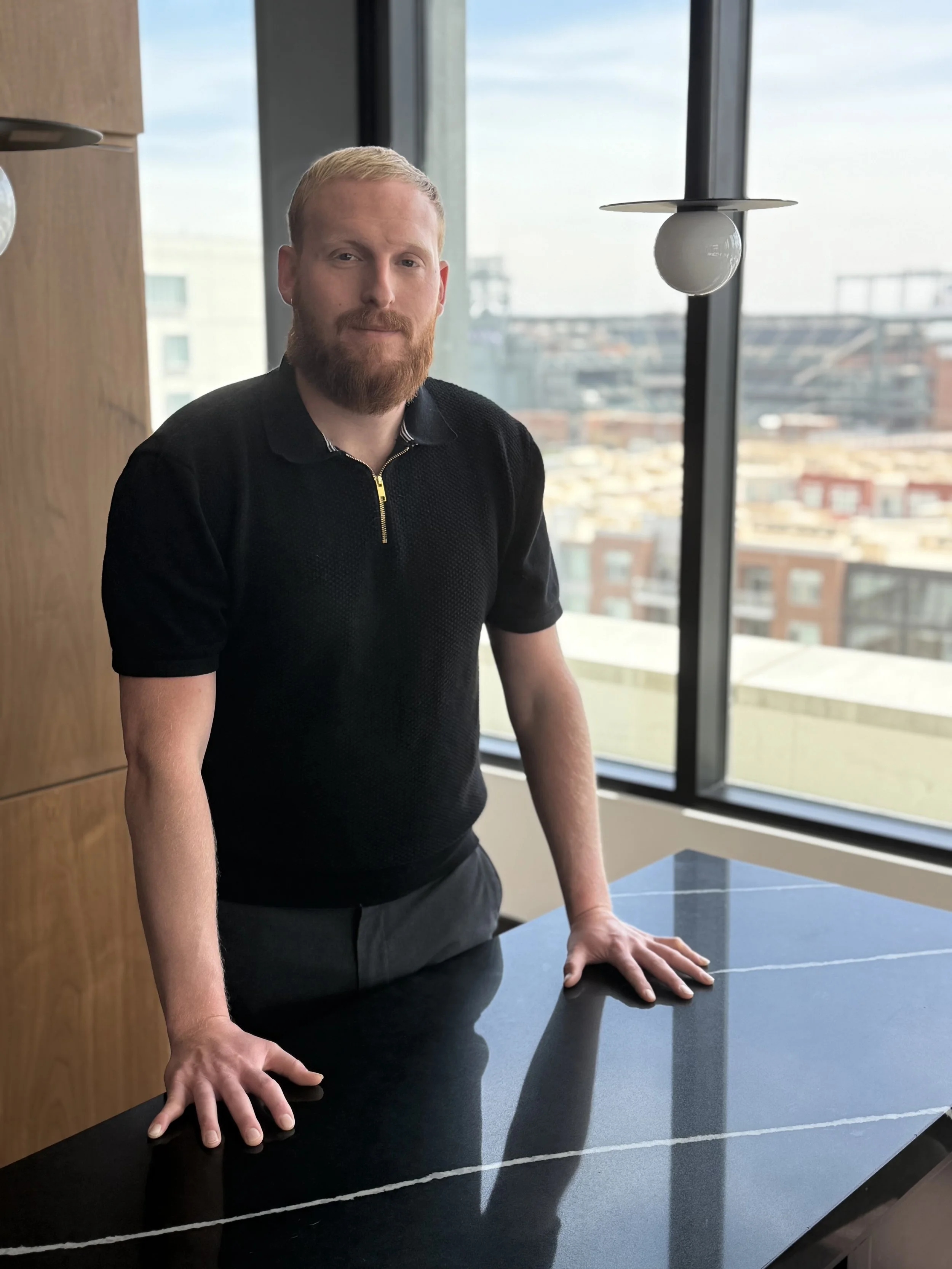 A man with red hair and a beard standing indoors at a table with his hands resting on its surface, near a large window and a modern ceiling light fixture.
