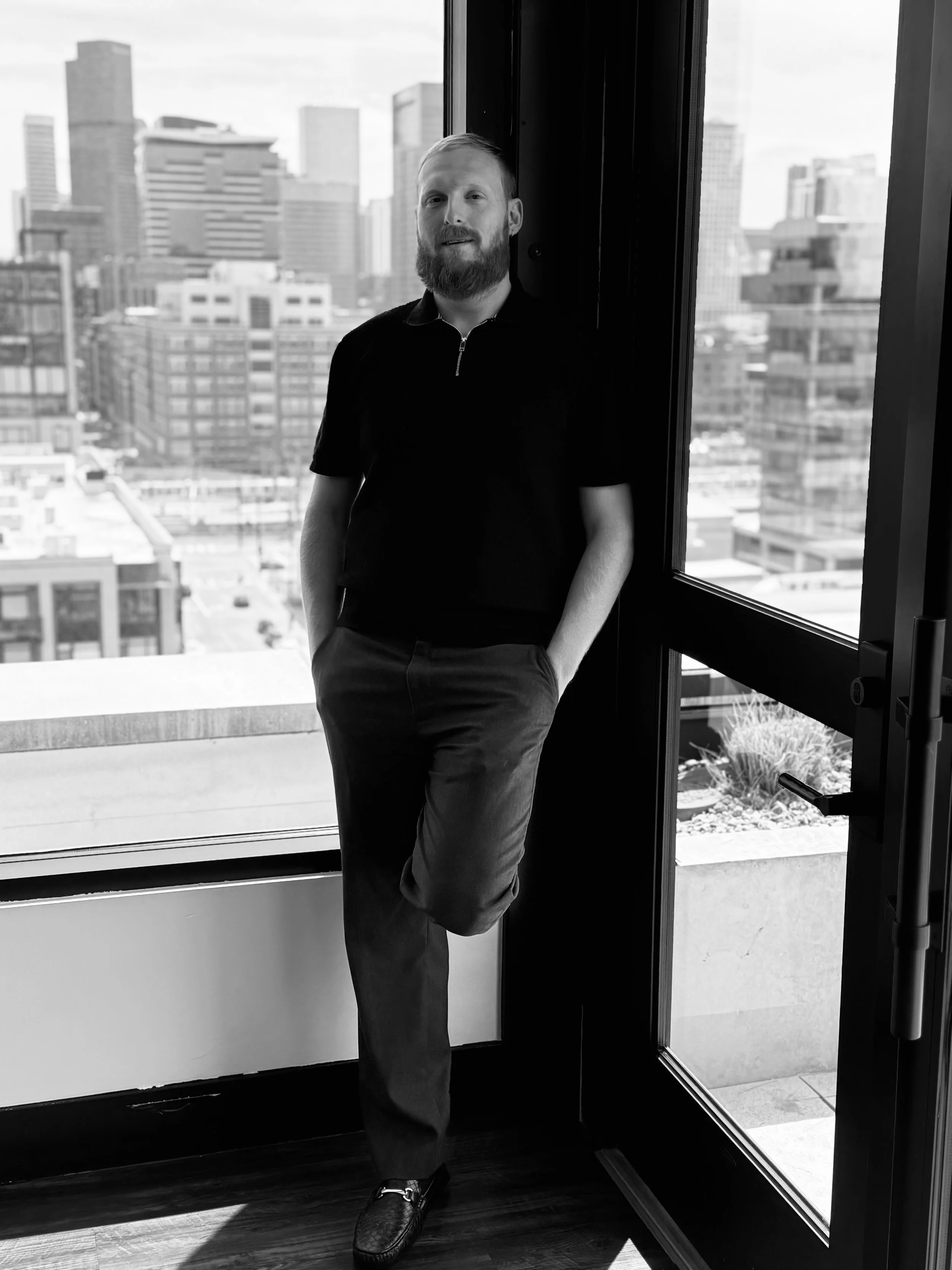 A man with a beard and short hair standing indoors near a large window with a city skyline in the background, wearing a dark polo shirt, pants, and loafers.