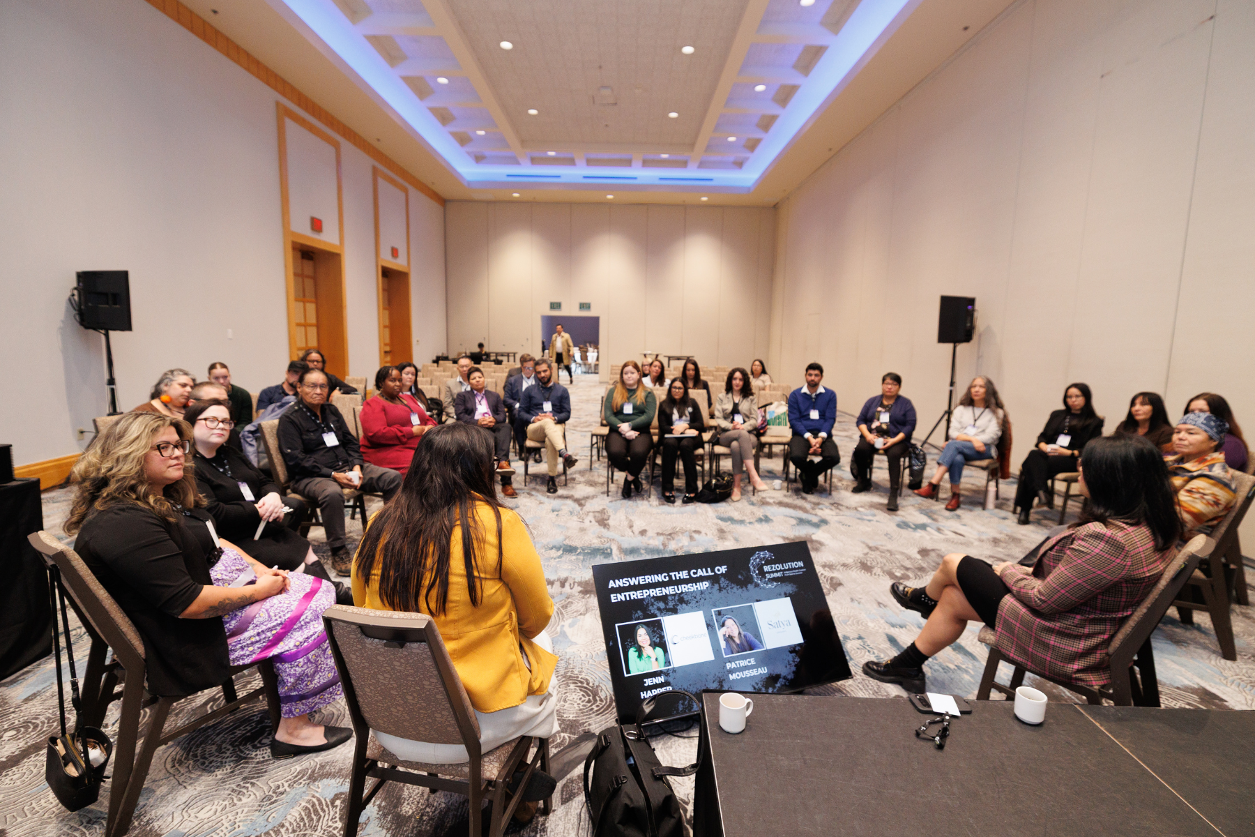 A diverse group of people sitting in a circle in a conference room, engaged in a discussion. In the foreground, a woman in a pink and orange striped top is speaking, with a large screen displaying their names and a presentation behind her.