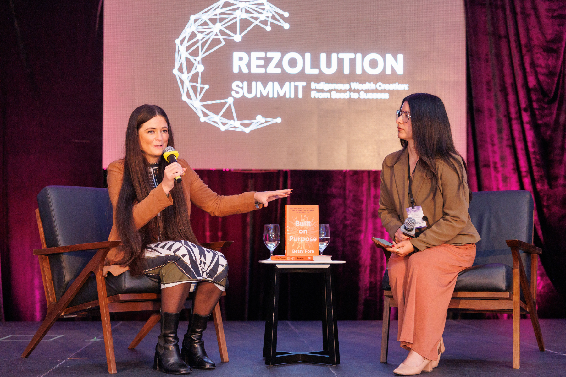 Two women on stage at the Rezolution Summit. One woman is speaking into a microphone while the other listens and holds a microphone. A book titled 'Built on Purpose' by Betsy Fore is placed on a small table between them, with two glasses of water.