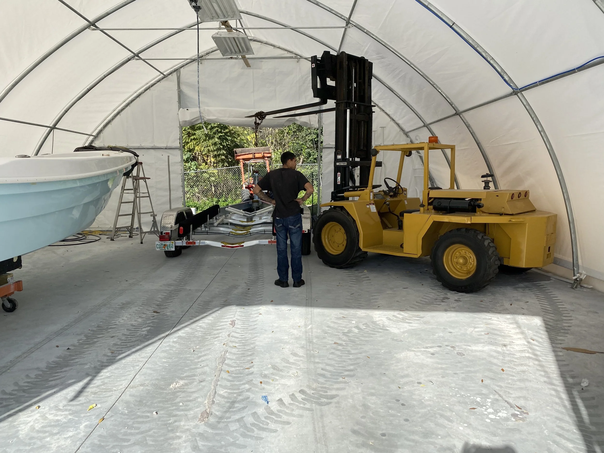 A man standing inside a large indoor storage tent, near a yellow forklift and a boat trailer with a boat on it, with a ladder and various equipment around.