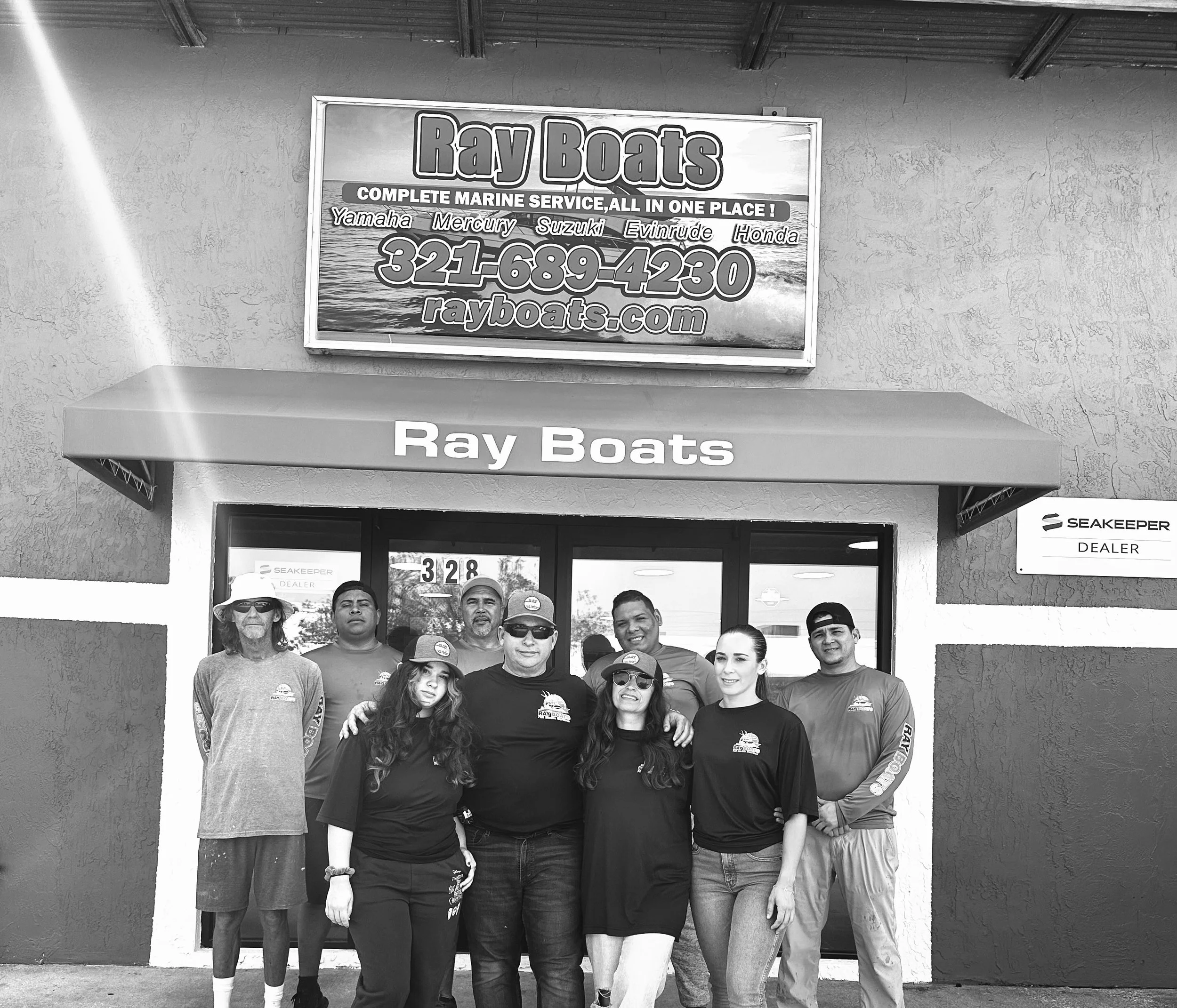 Group of eight people standing in front of Ray Boats dealership, smiling at the camera, with a sign above the entrance and a sign to the right.