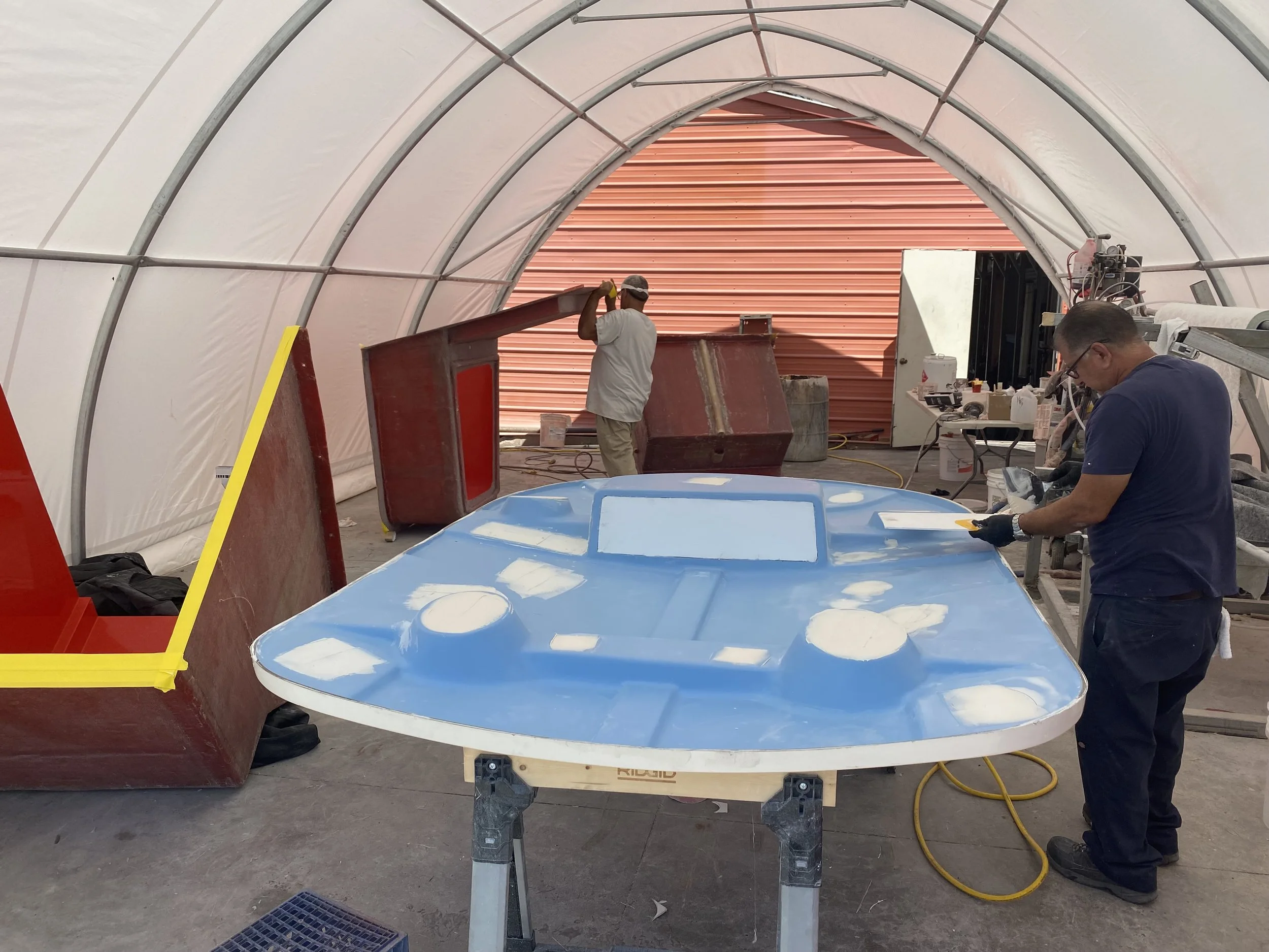Two workers in a workshop building a model of a blue and white spacecraft or aircraft. The space is a large, covered area with various tools and materials around.