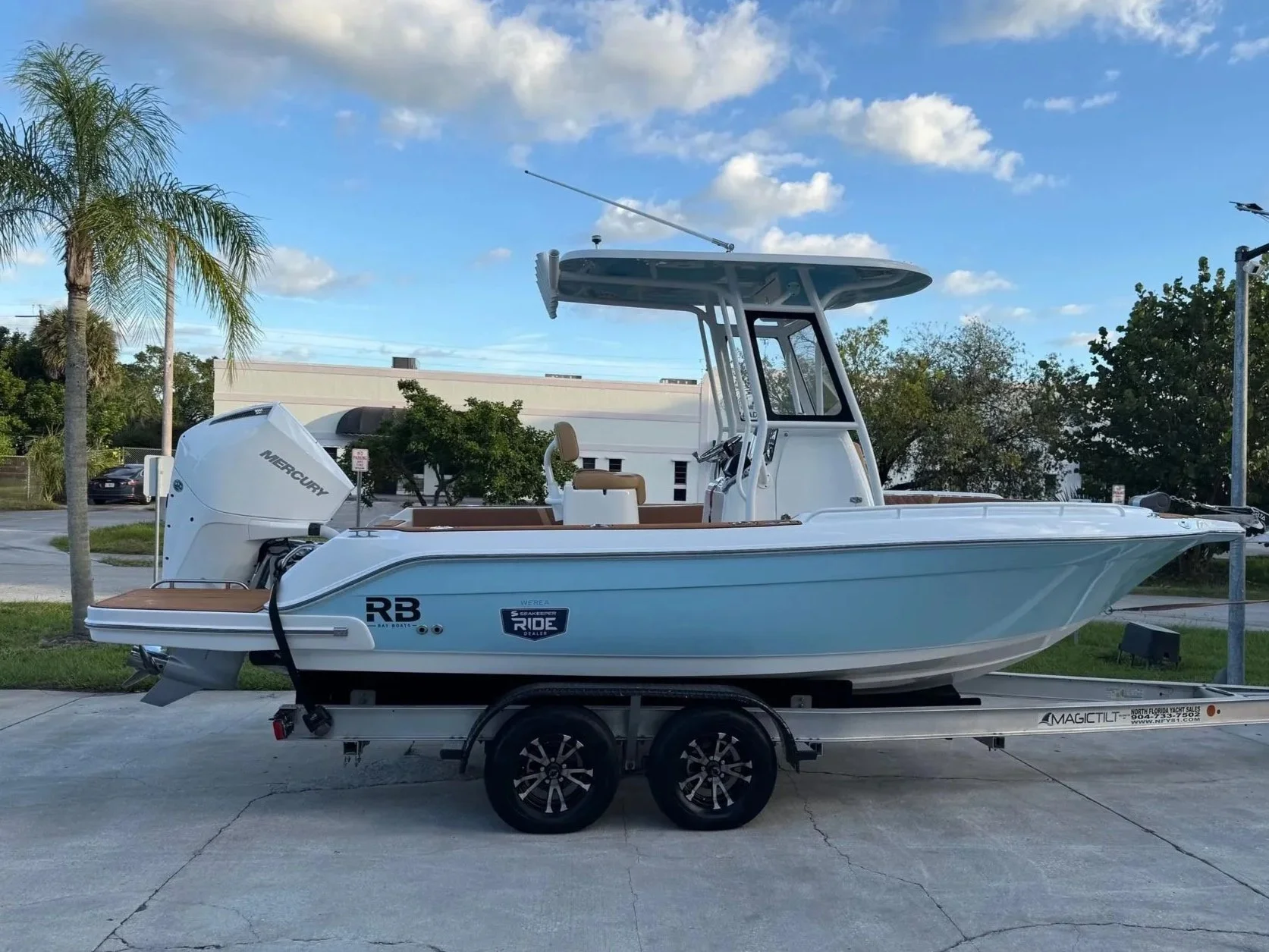 A light blue and white boat on a double-axle trailer parked on a concrete surface with palm trees and buildings in the background.