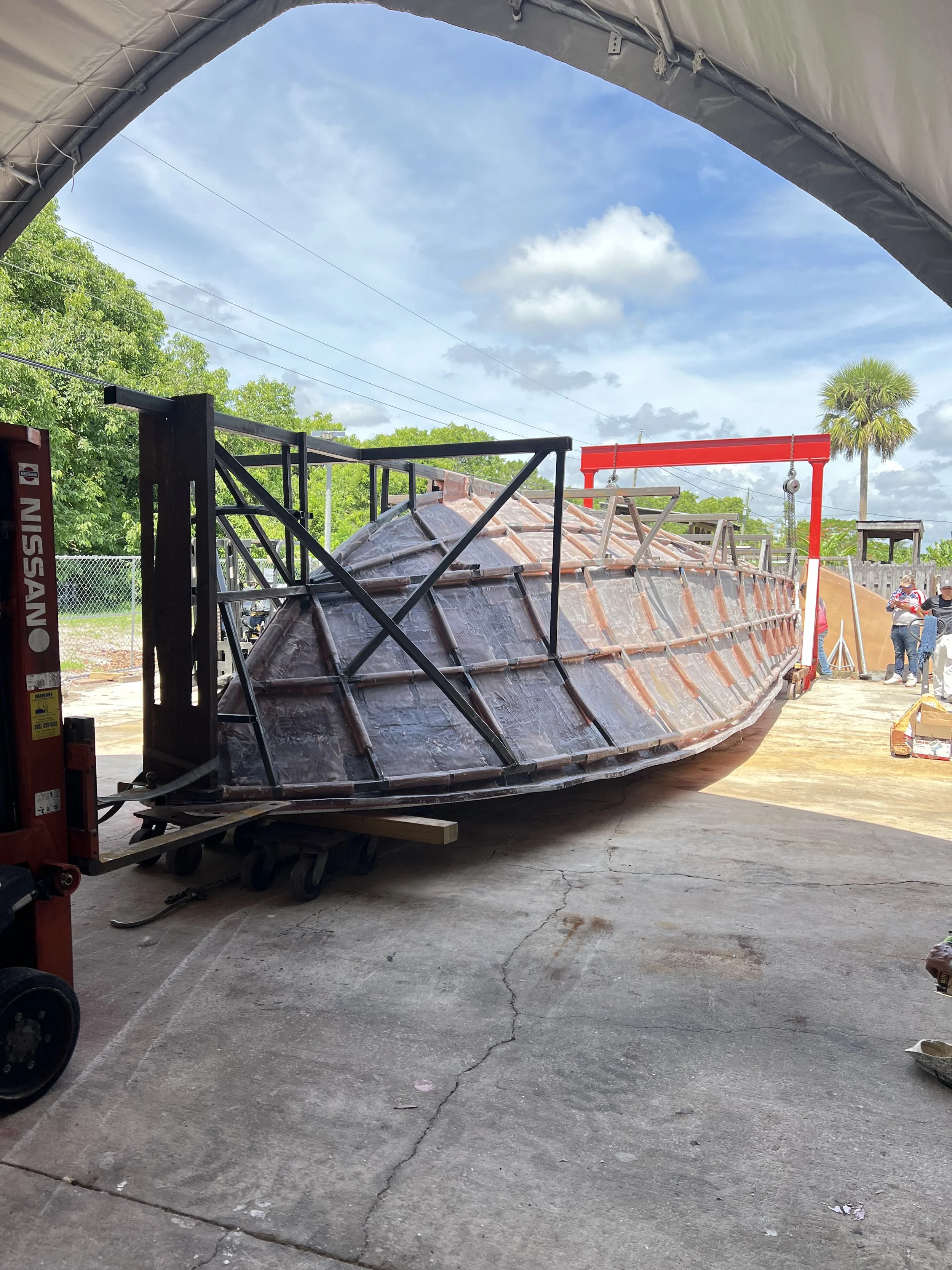 A boat under construction or repair, supported by a metal frame, inside a covered building. Several workers are visible in the background.