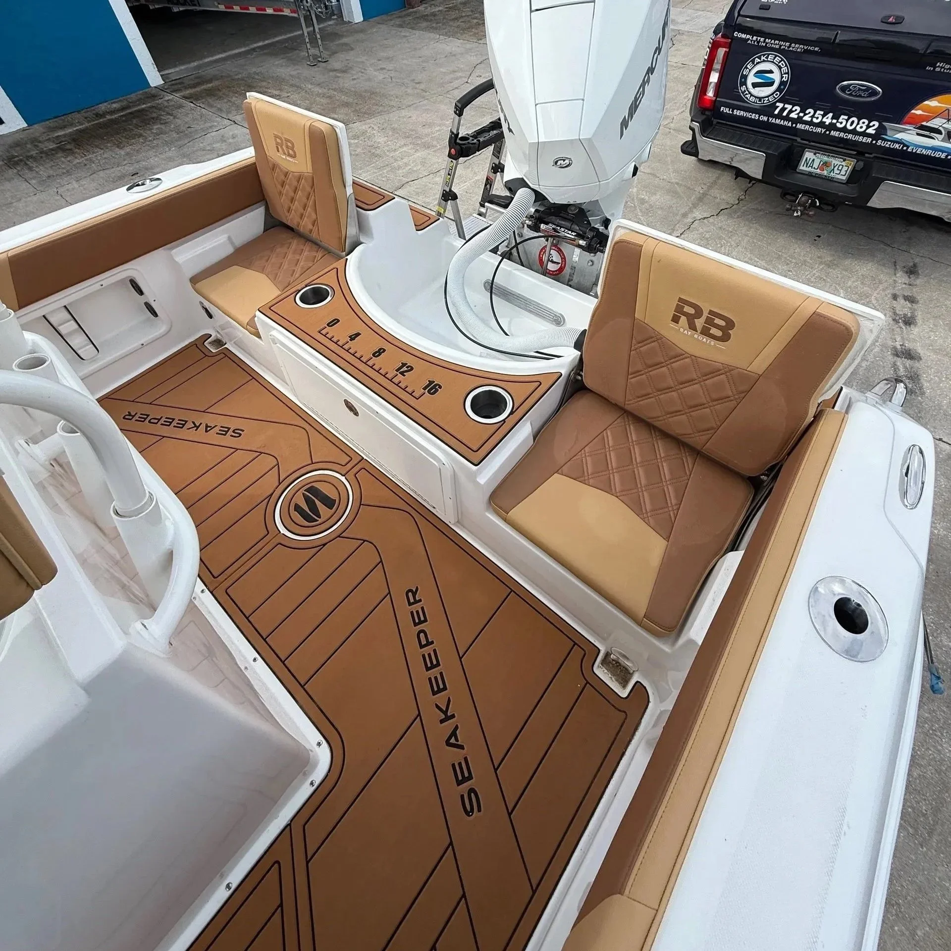 Inside a boat with tan cushioned seats, a brown and tan mat with the words "SEAKEEPER" and the logo, and a white console with a mounted outboard engine.