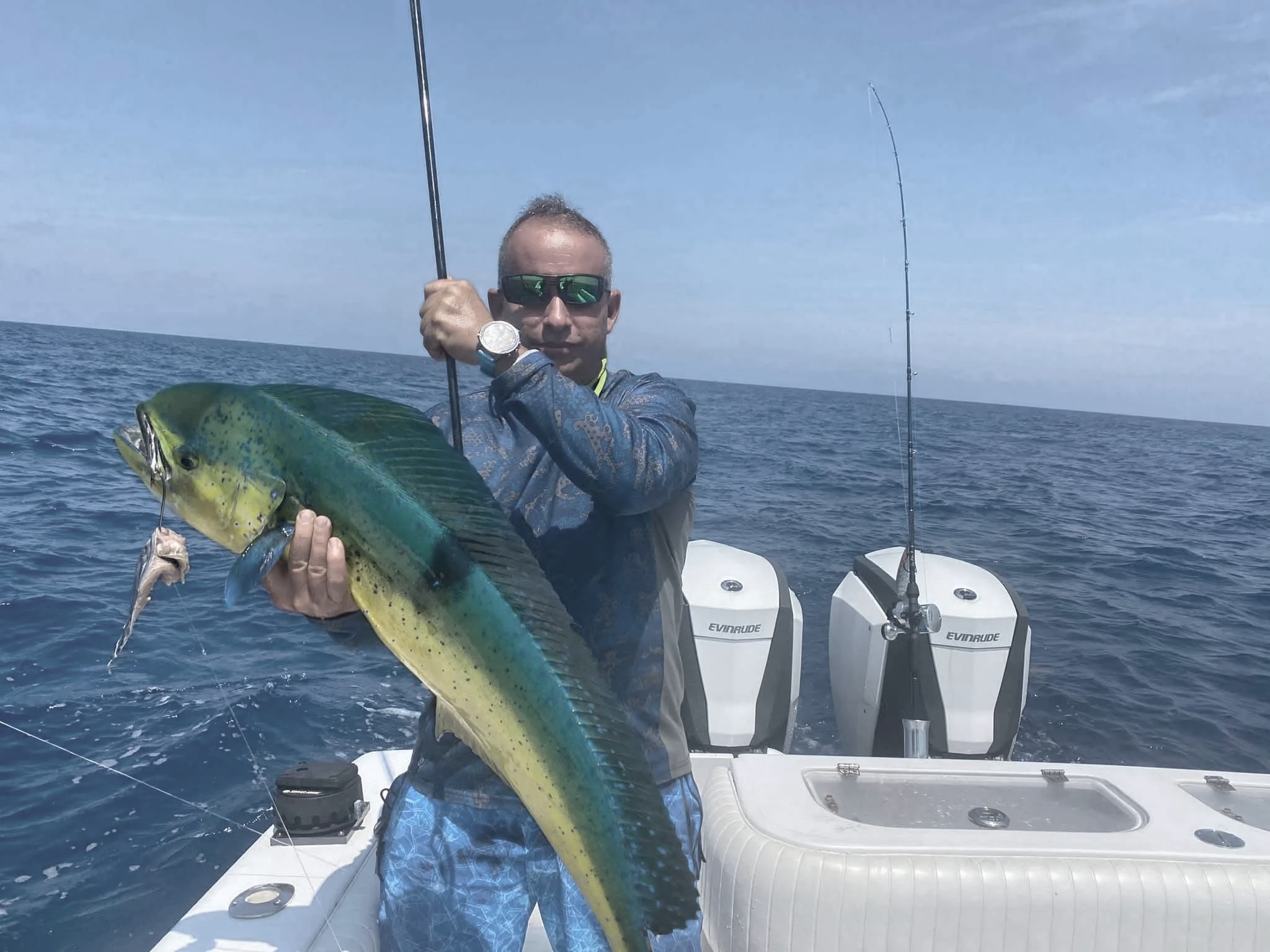 Man wearing sunglasses and a watch holding a large fish on a boat at sea, with fishing rods and motors in the background.