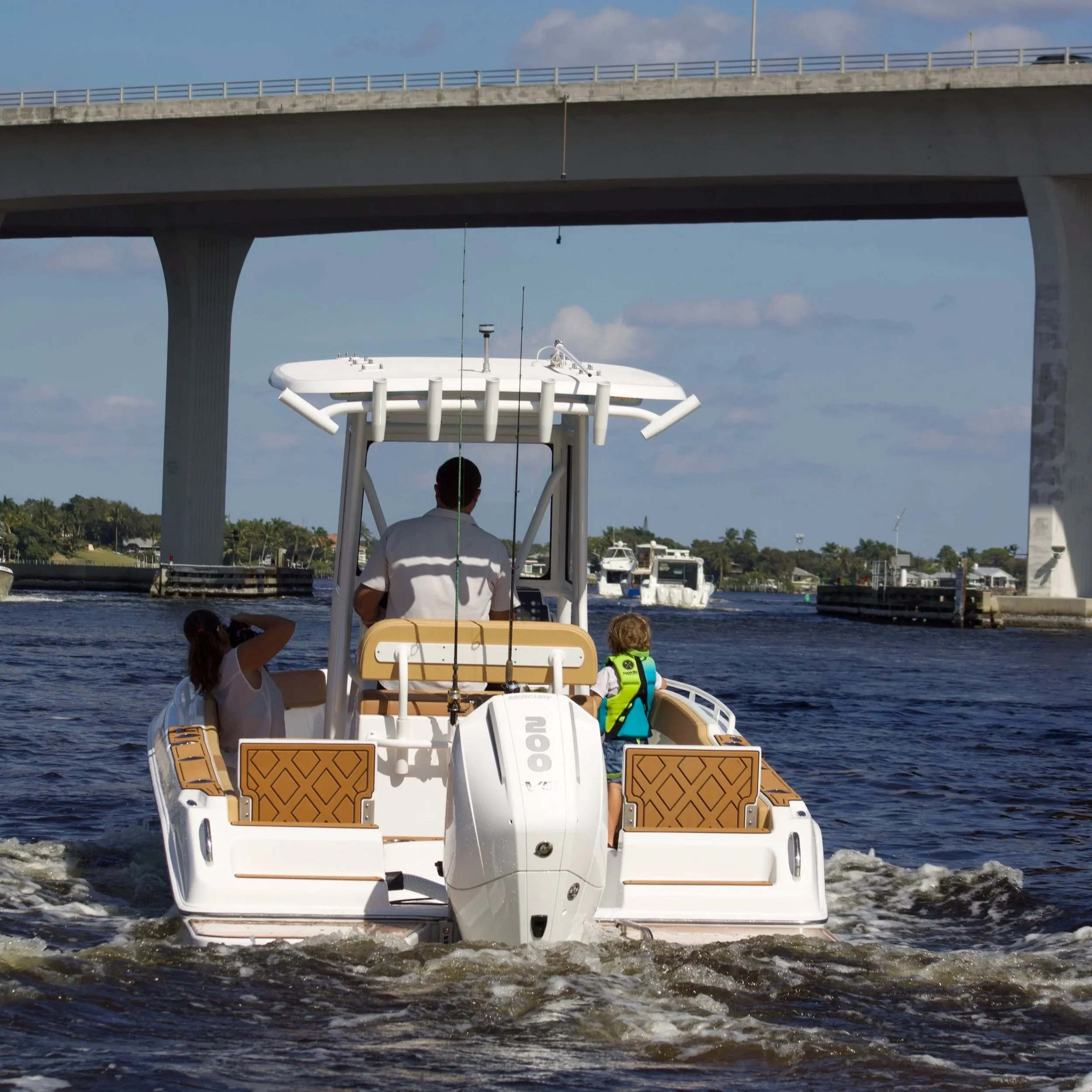 A white motorboat with two passengers, a man and a woman, moving under a bridge on a body of water. The woman wears a life vest and the man is steering the boat. In the background, there are other boats and greenery along the shoreline.