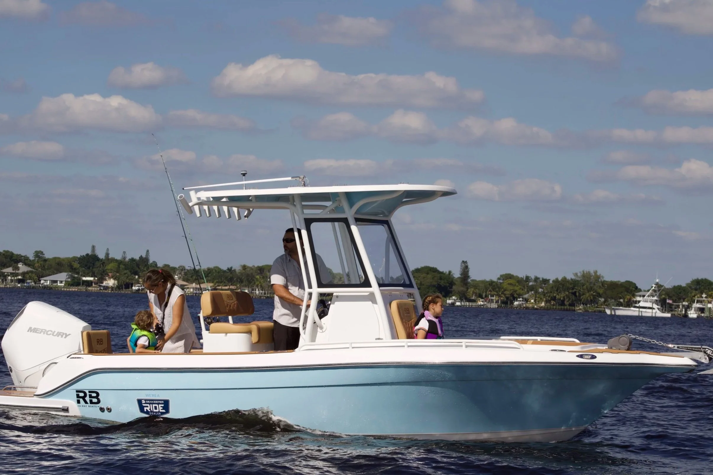 A white and tan boat with two children and two adults on a lake with houses and trees in the background, under a partly cloudy sky.