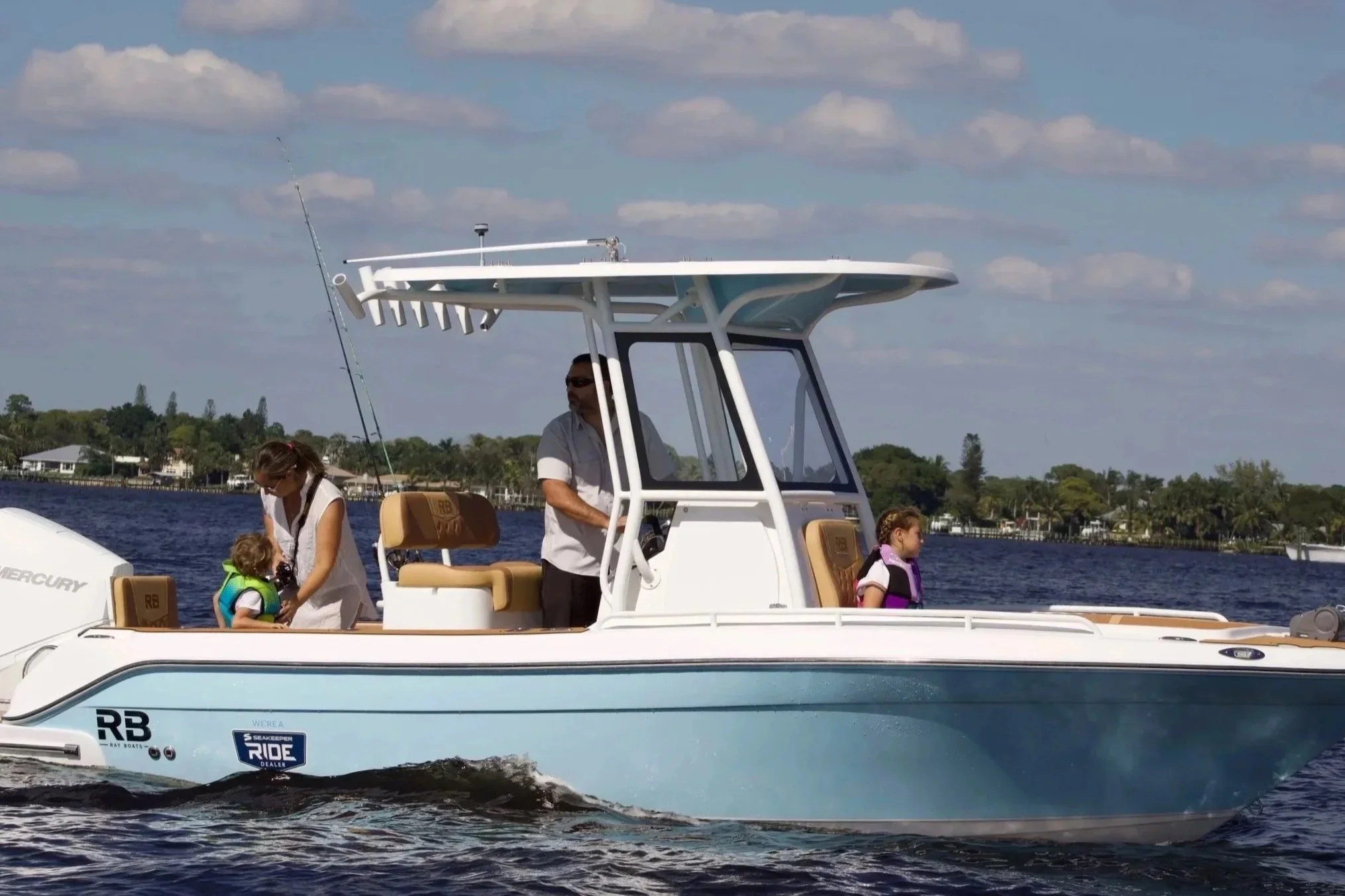 A family on a white motorboat with tan seating, sailing on a body of water with a shoreline and houses in the background. The boat has a central cabin and a fishing rod mounted on top. Two children are sitting on the boat with life jackets, while an adult woman is adjusting something on one of the children, and an adult man is standing at the wheel.
