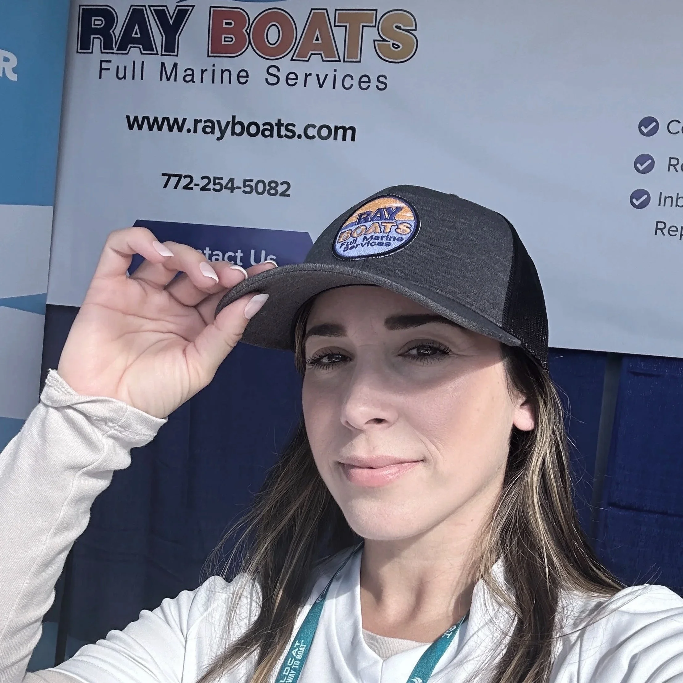 Woman wearing a gray trucker hat with the logo of Ray Boats, standing in front of a sign for Ray Boats Full Marine Services with contact information and website.