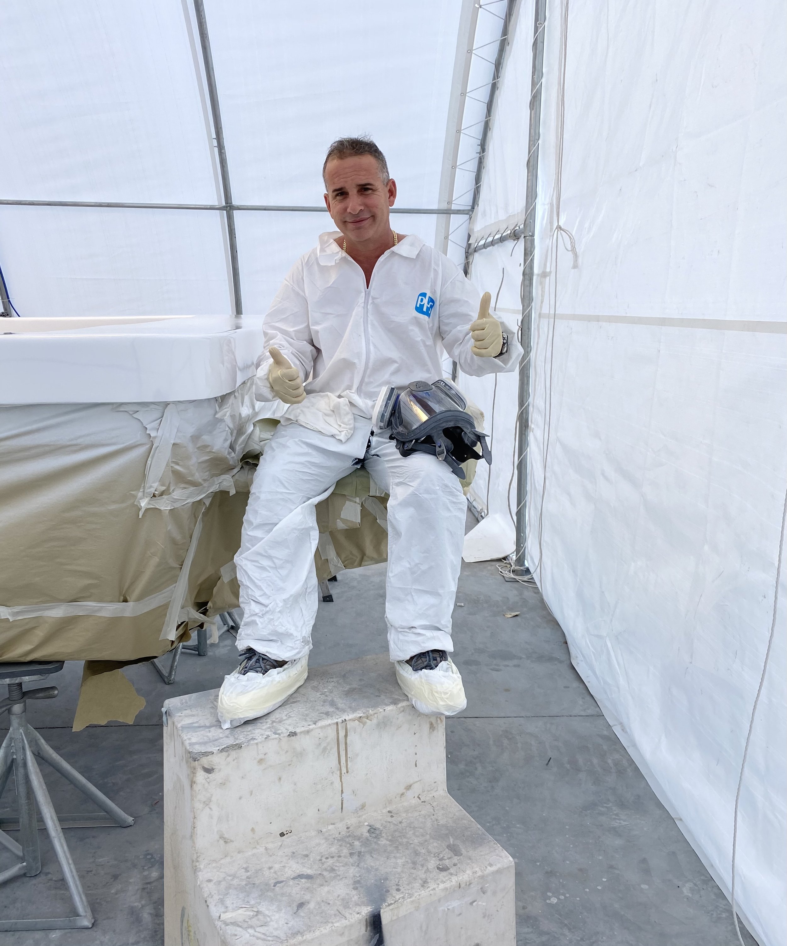 Man dressed in white protective suit giving a thumbs-up inside a white tent with construction or laboratory equipment visible.