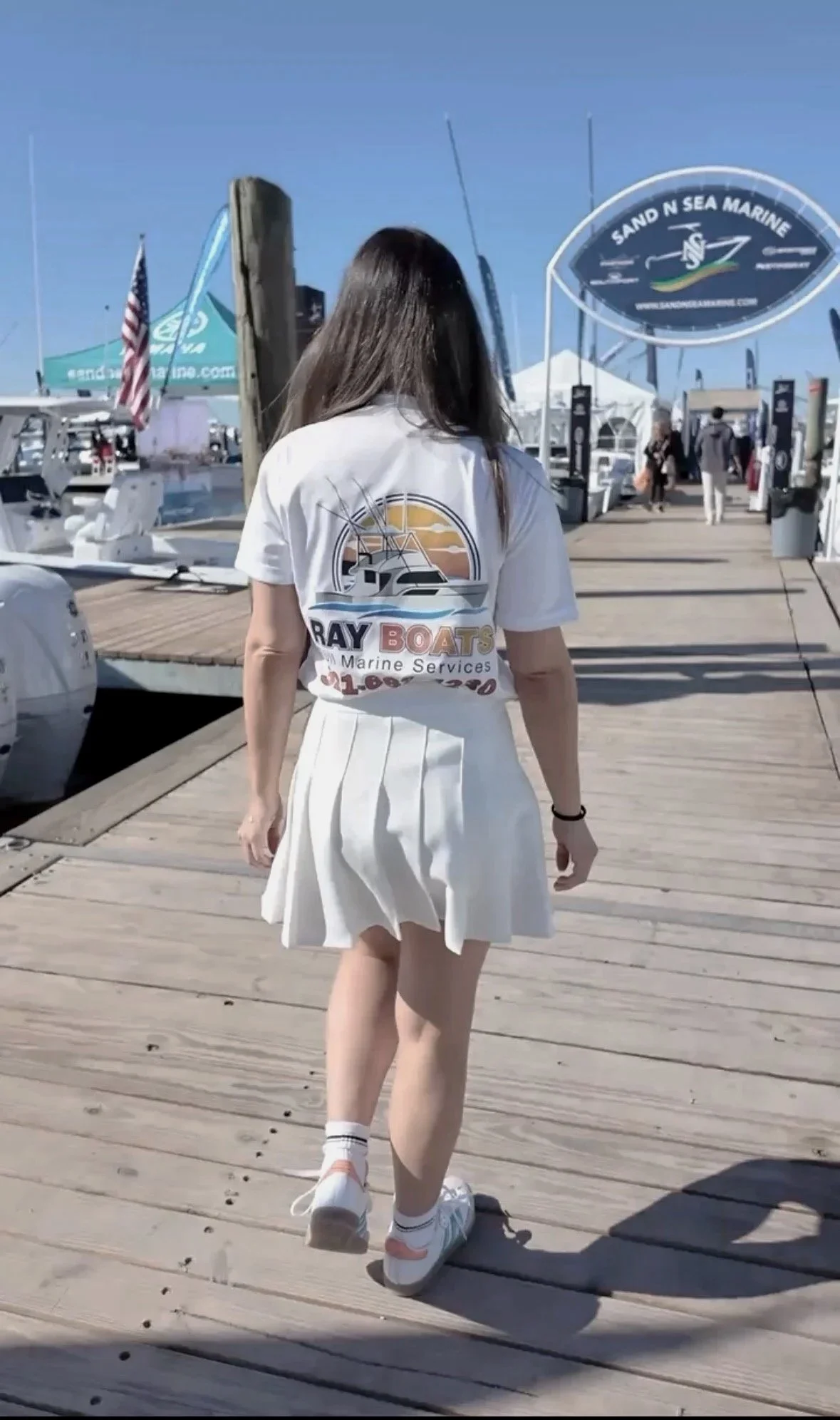 A woman walking on a wooden dock at a marina, wearing a white t-shirt with a boat logo and white pleated skirt, with boats and marina signs in the background on clear, sunny day.