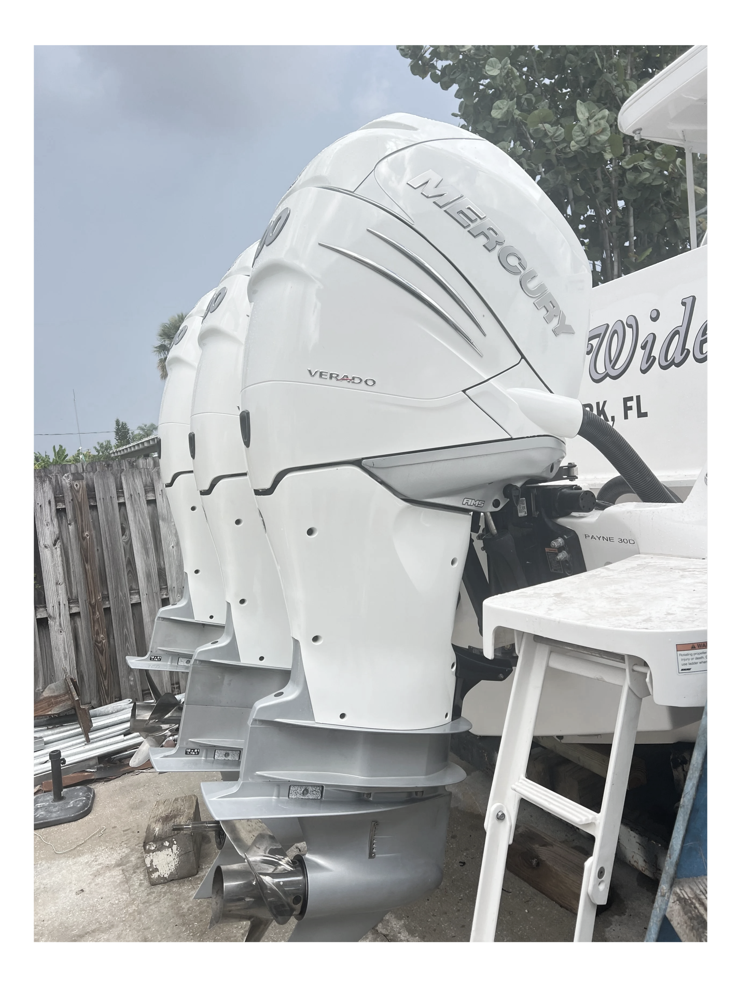 Close-up of three white Mercury Verado outboard motors attached to a boat, with a wooden fence and trees in the background.
