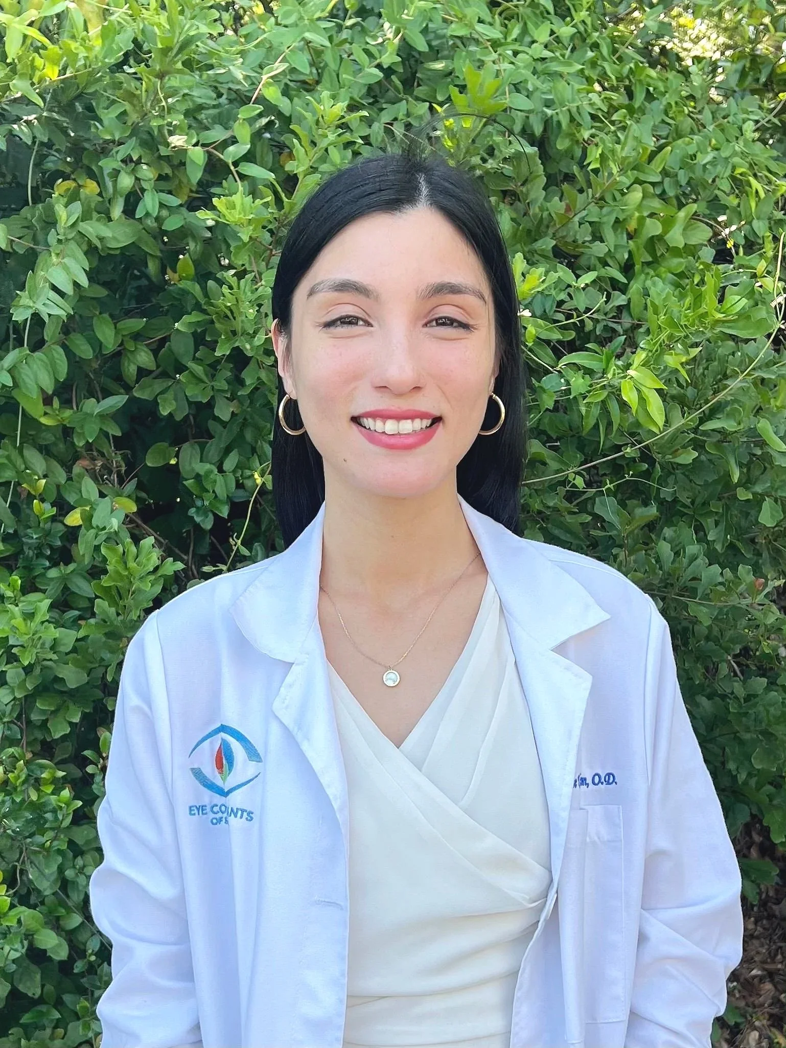 A woman with black hair and hoop earrings smiling outdoors against a background of green foliage, wearing a white lab coat with logos and text