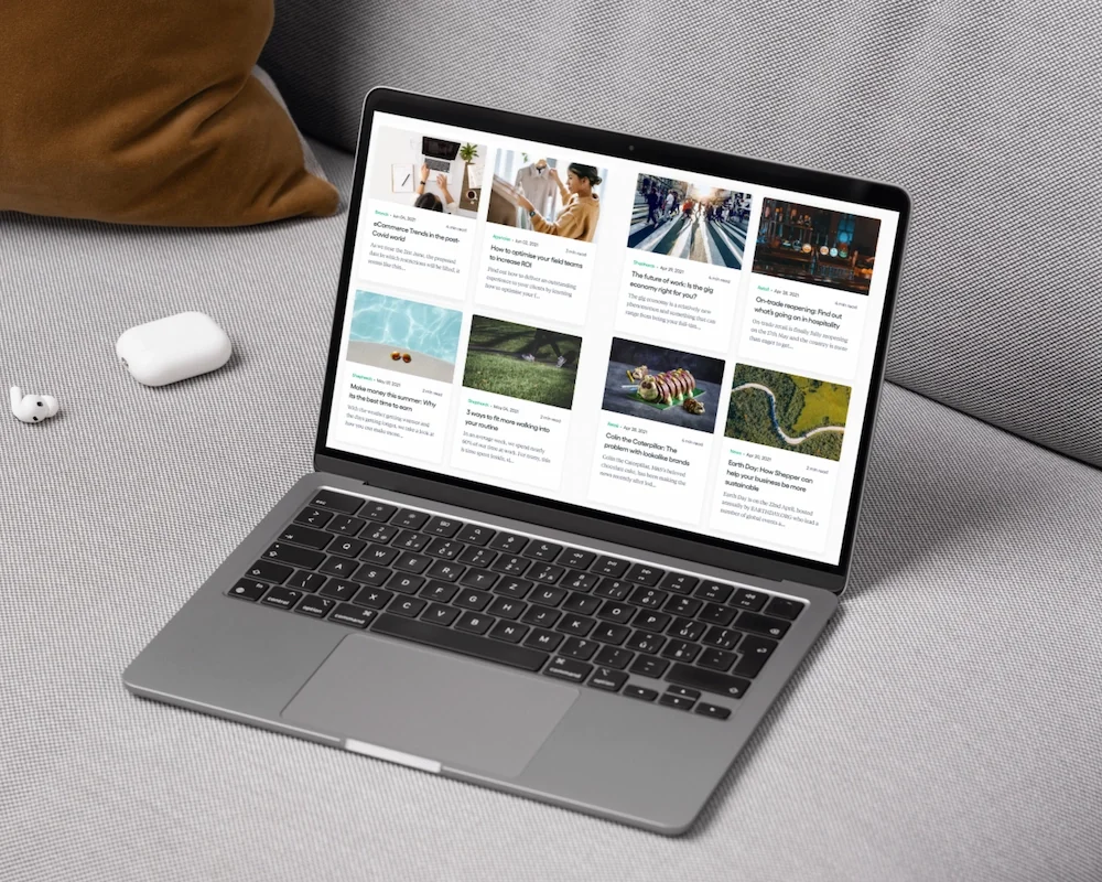 Open laptop on a gray surface displaying a webpage with articles and images. Near the laptop are wireless earbuds and a smartphone with a brown case. Part of a person's arm in a brown sofa is visible.