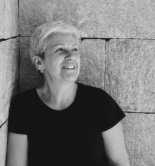Fotografia di Lara Galanti, l'architetta proprietaria di Filorosso design, con capelli corti biondi, che sorride, indossa una maglietta nera e si trova accanto a un muro di pietra