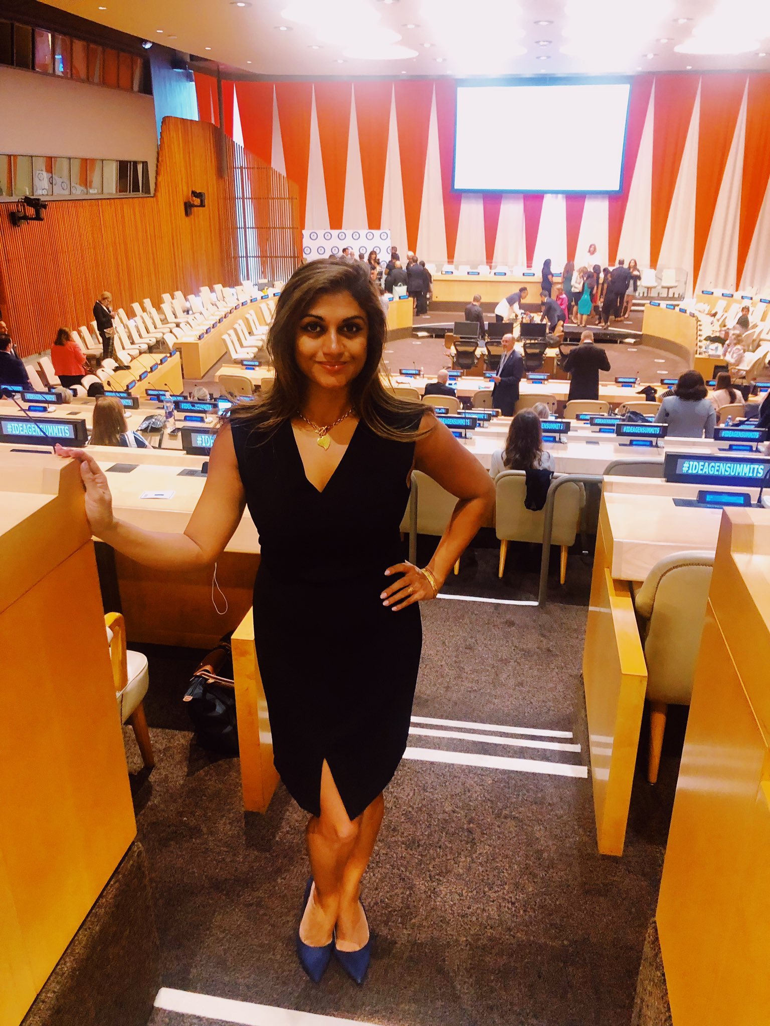 A woman in a black dress and heels standing inside a conference or summit venue with a large stage and many attendees behind her.