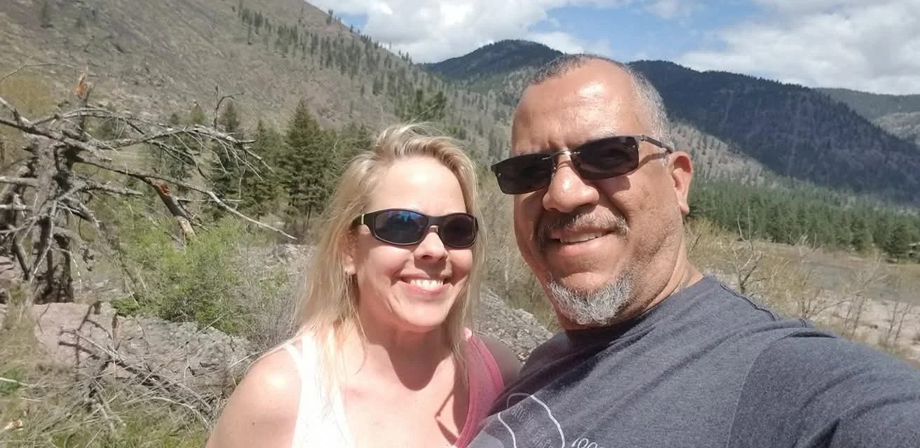 A smiling couple wearing sunglasses taking a selfie outdoors in a mountainous area with trees and overcast sky.