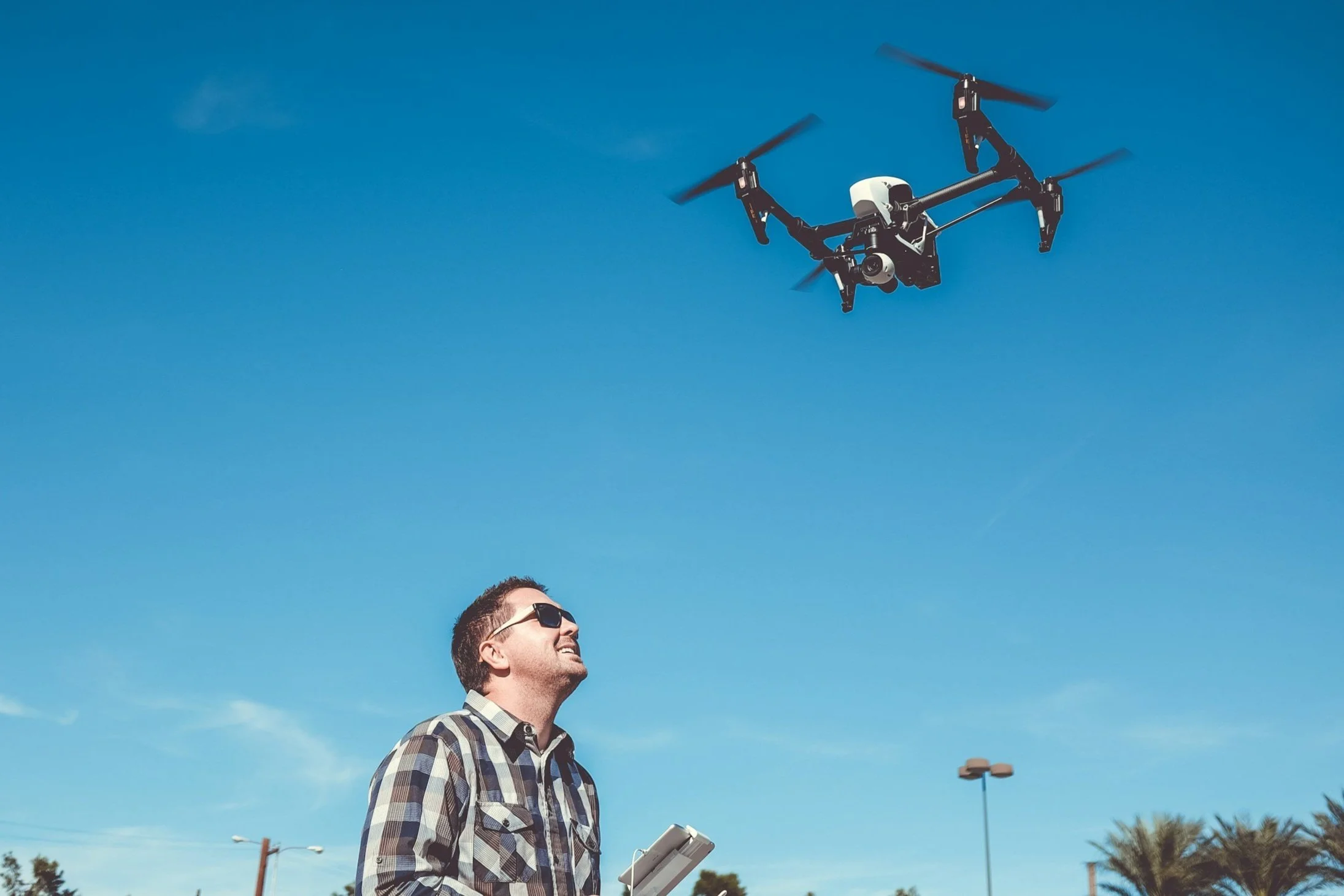 A man with sunglasses flying a drone in a clear blue sky, holding a remote control.