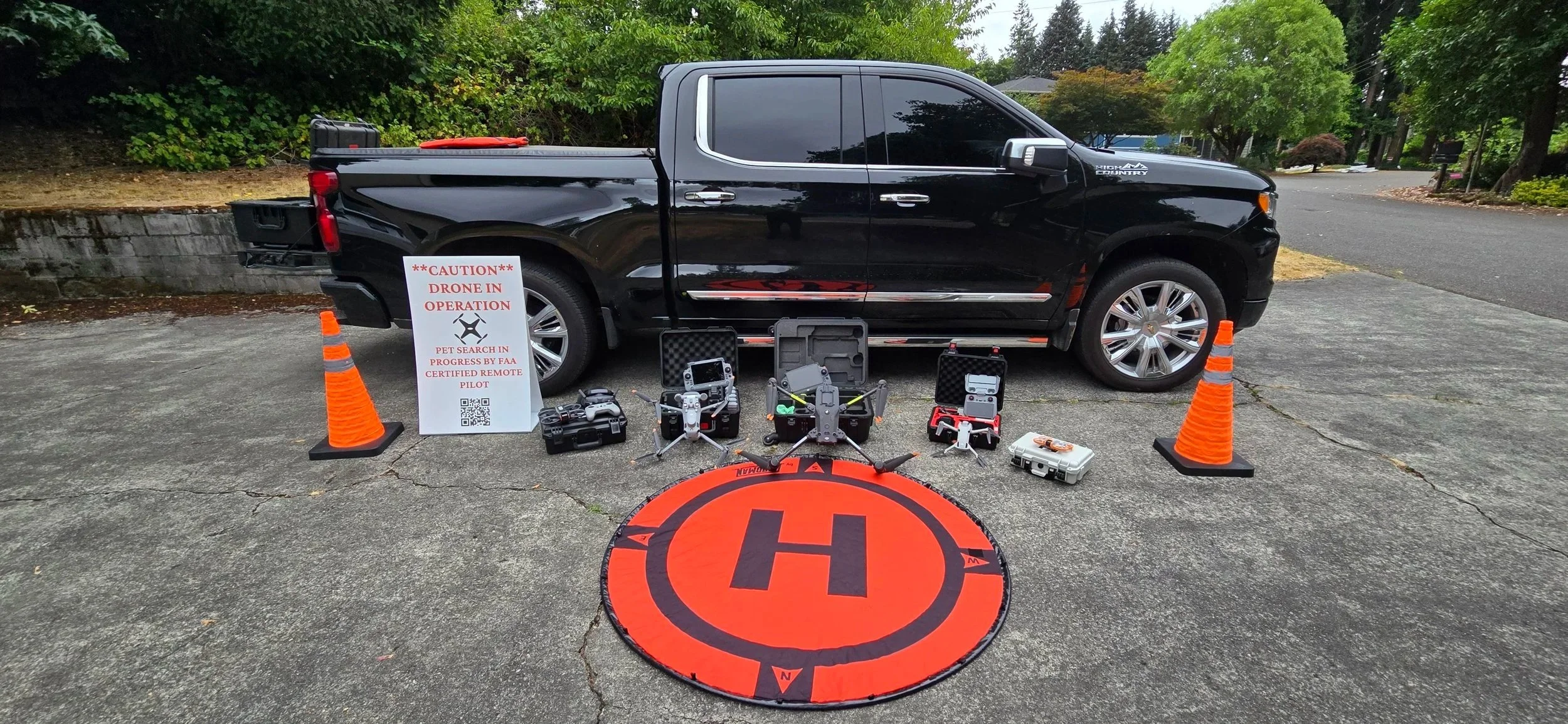 Black pickup truck parked on the street with orange traffic cones on each side. In front of the truck, there are multiple drones, remote controls, and equipment cases. A sign on the left reads '**CAUTION** DRONE IN OPERATION PET SEARCH IN PROGRESS BY FAA CERTIFIED REMOTE PILOT' with a QR code below. An orange helicopter landing pad with a large 'H' symbol is on the ground.