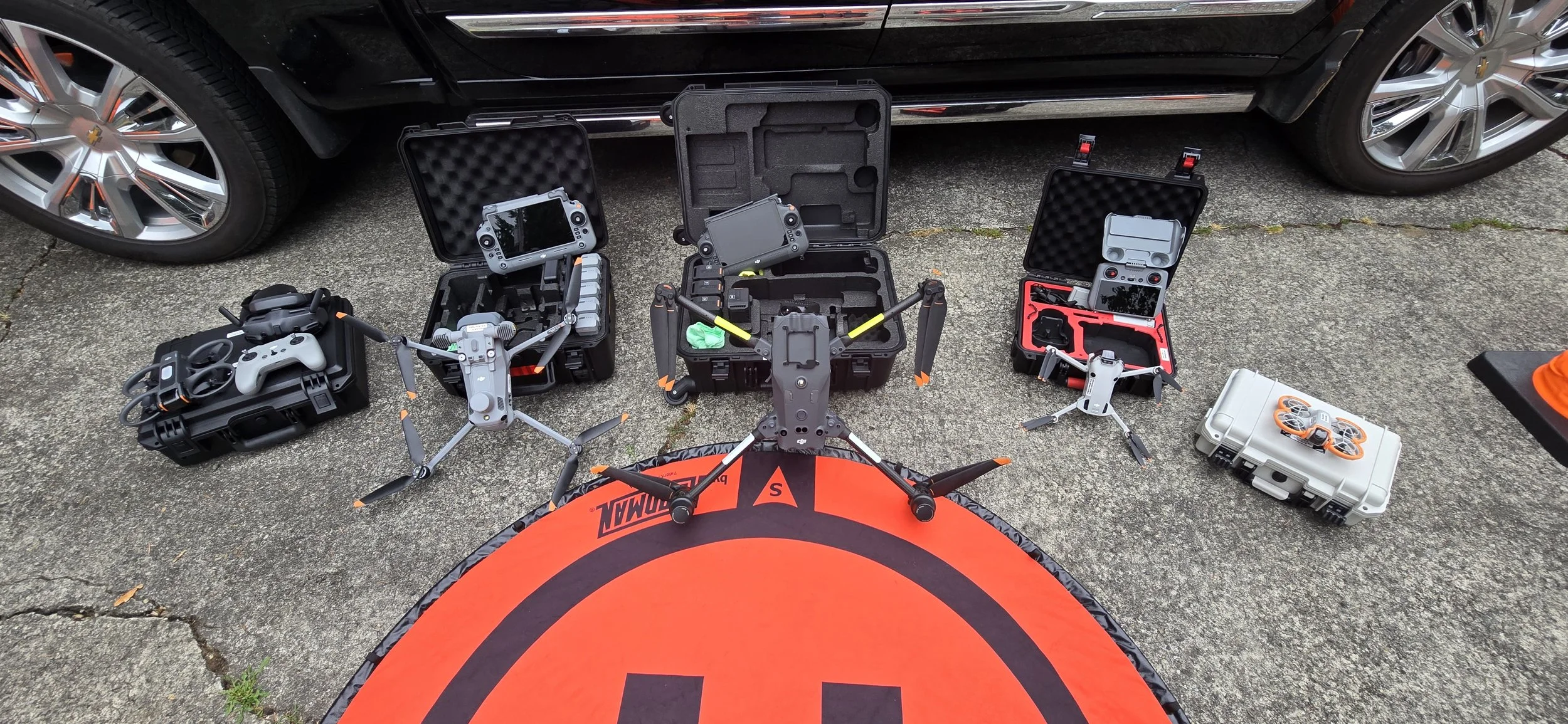 Drones, remote controls, and equipment cases arranged on a concrete driveway, with cars parked in the background.