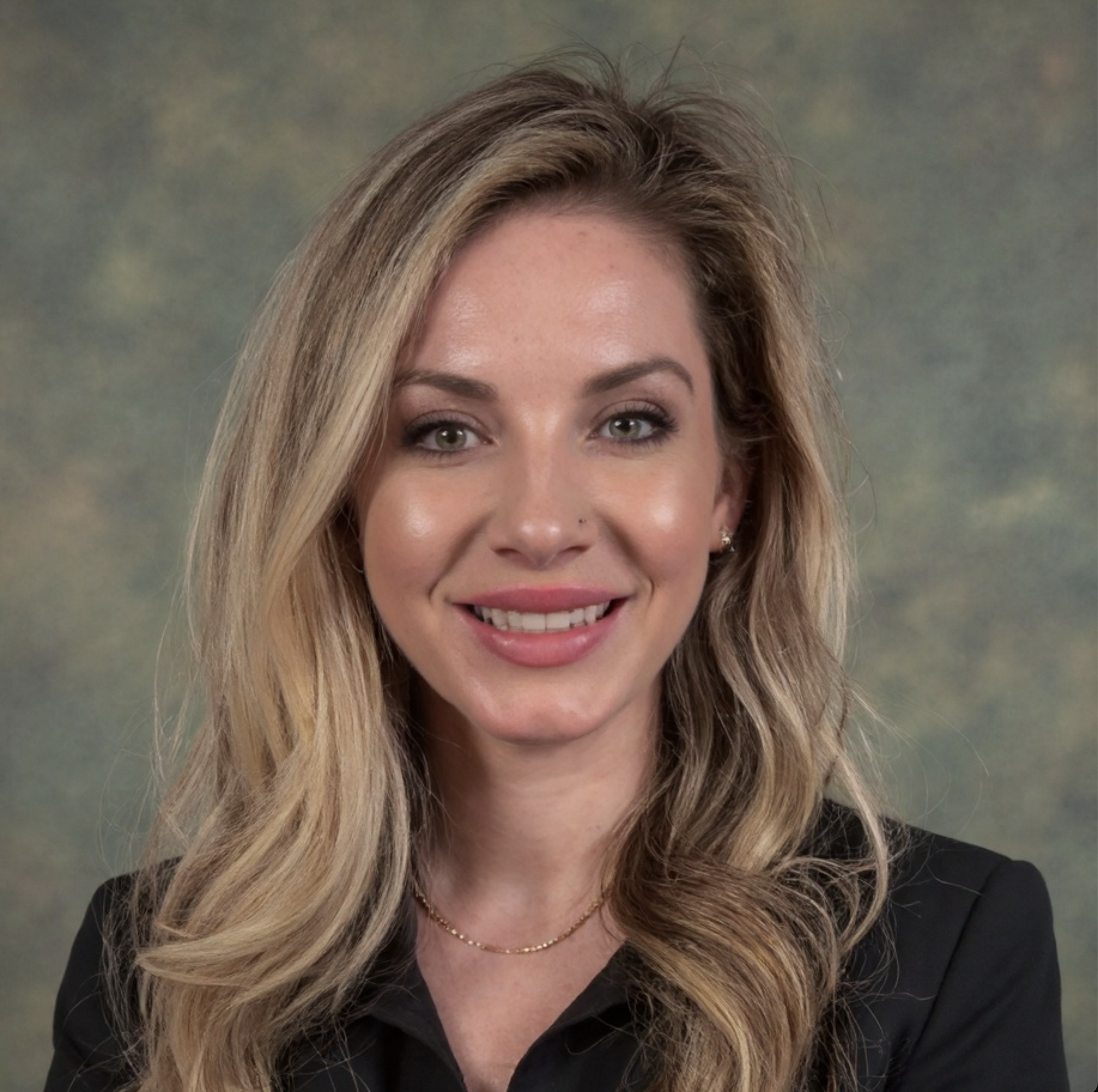 Portrait of a woman with blonde wavy hair, light skin, wearing a black blazer and a gold necklace, smiling against a muted background.