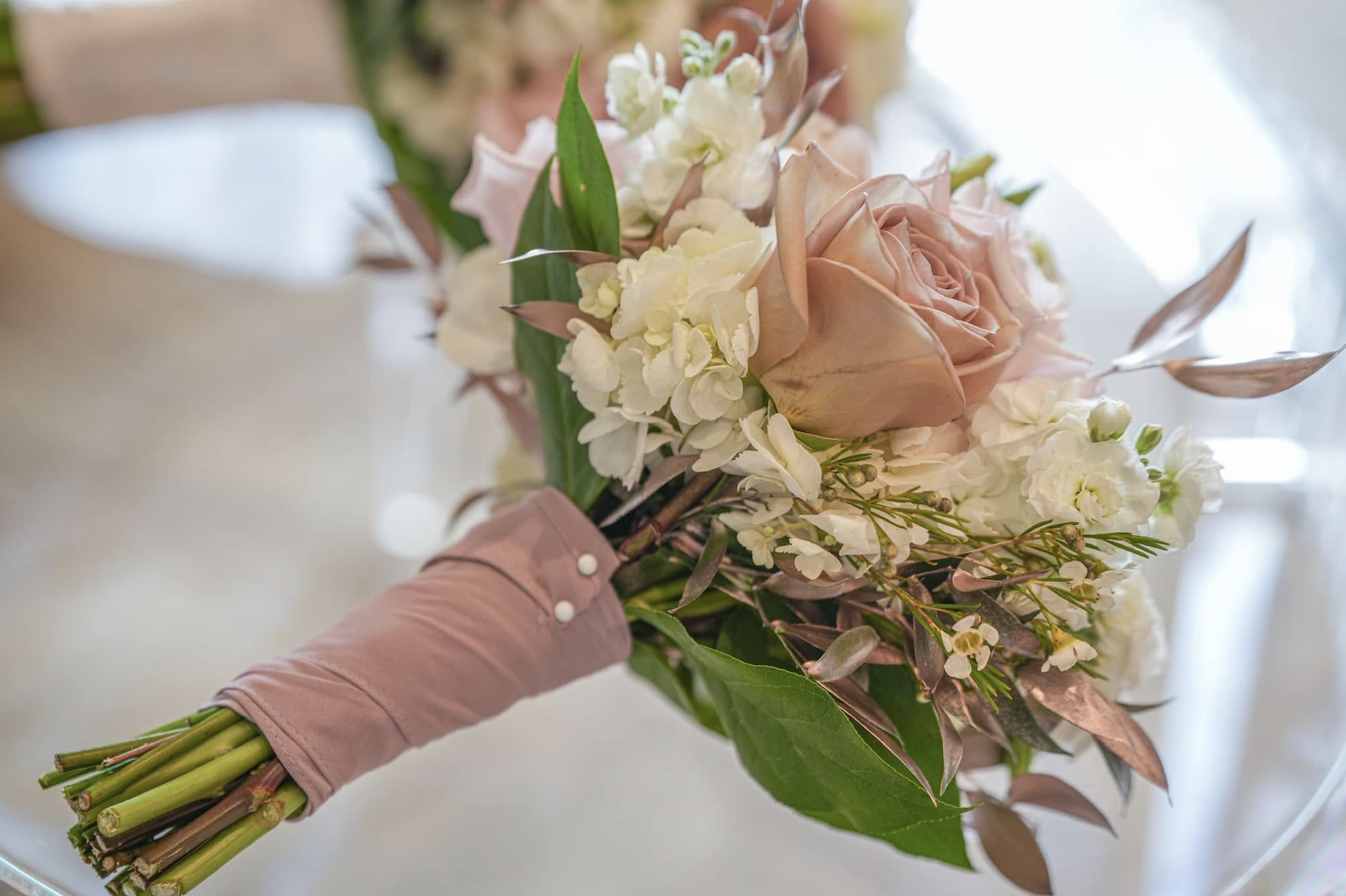 A person's hand holding a bouquet of flowers, including pink roses, white hydrangeas, and greenery. The person is wearing a long-sleeved pink shirt with pearl buttons on the cuff.