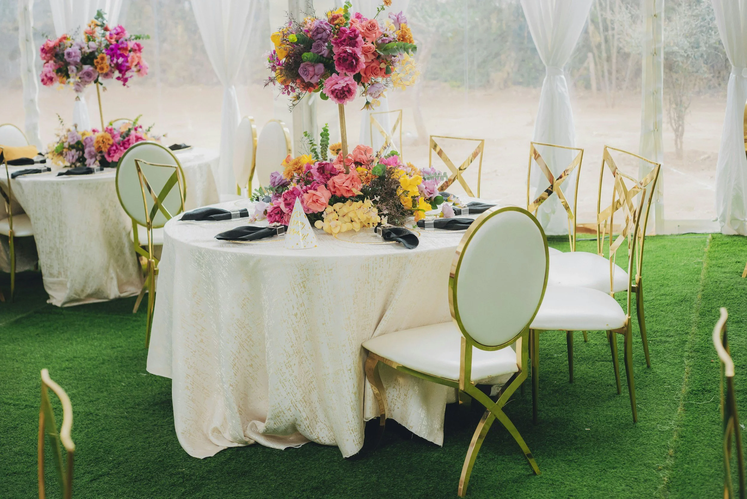 A decorated event table with a floral centerpiece on a white tablecloth, surrounded by white chairs with gold accents, set up outdoors on green grass with white draped curtains in the background.