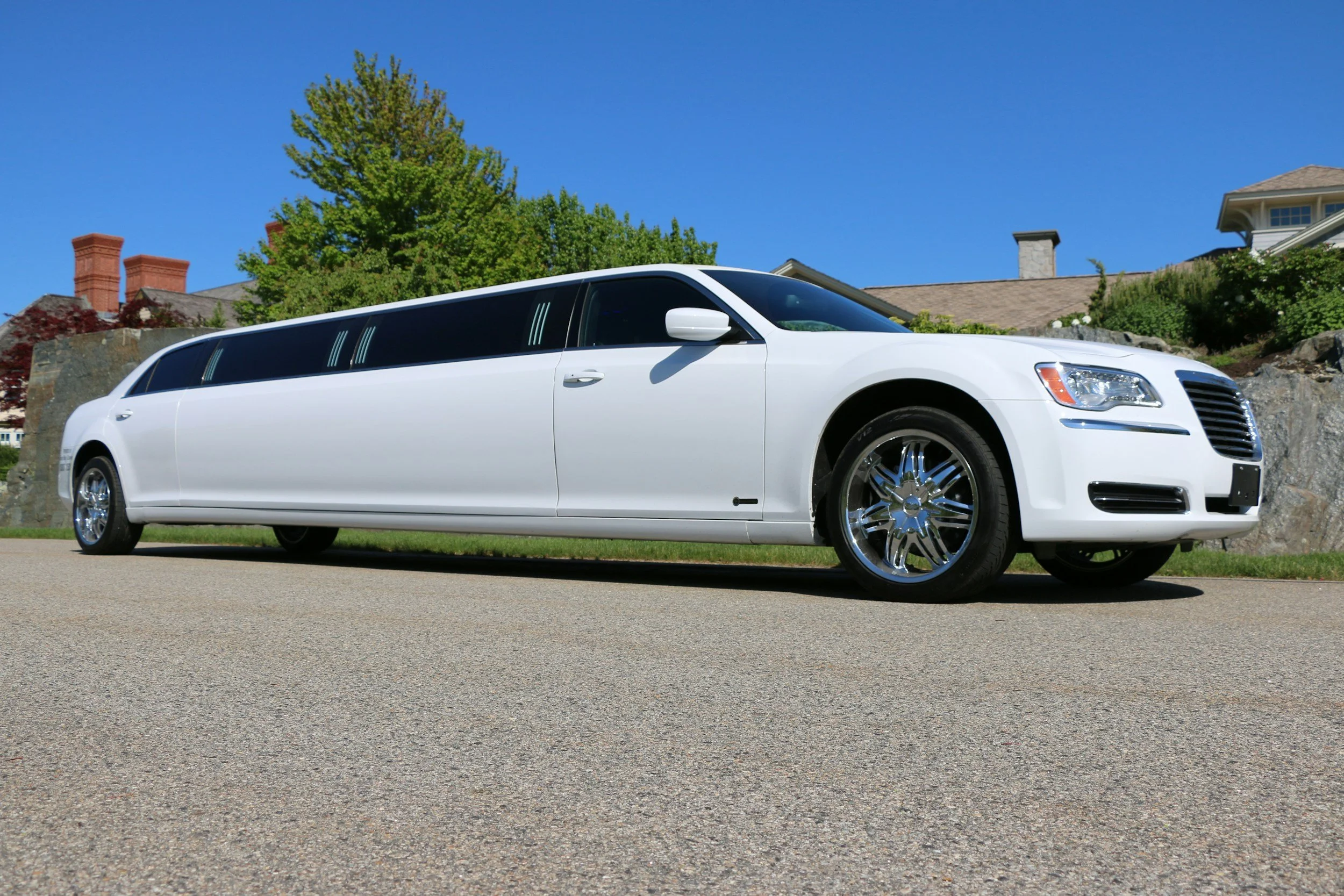 A white limo parked on the street in front of houses with trees and a blue sky in the background.