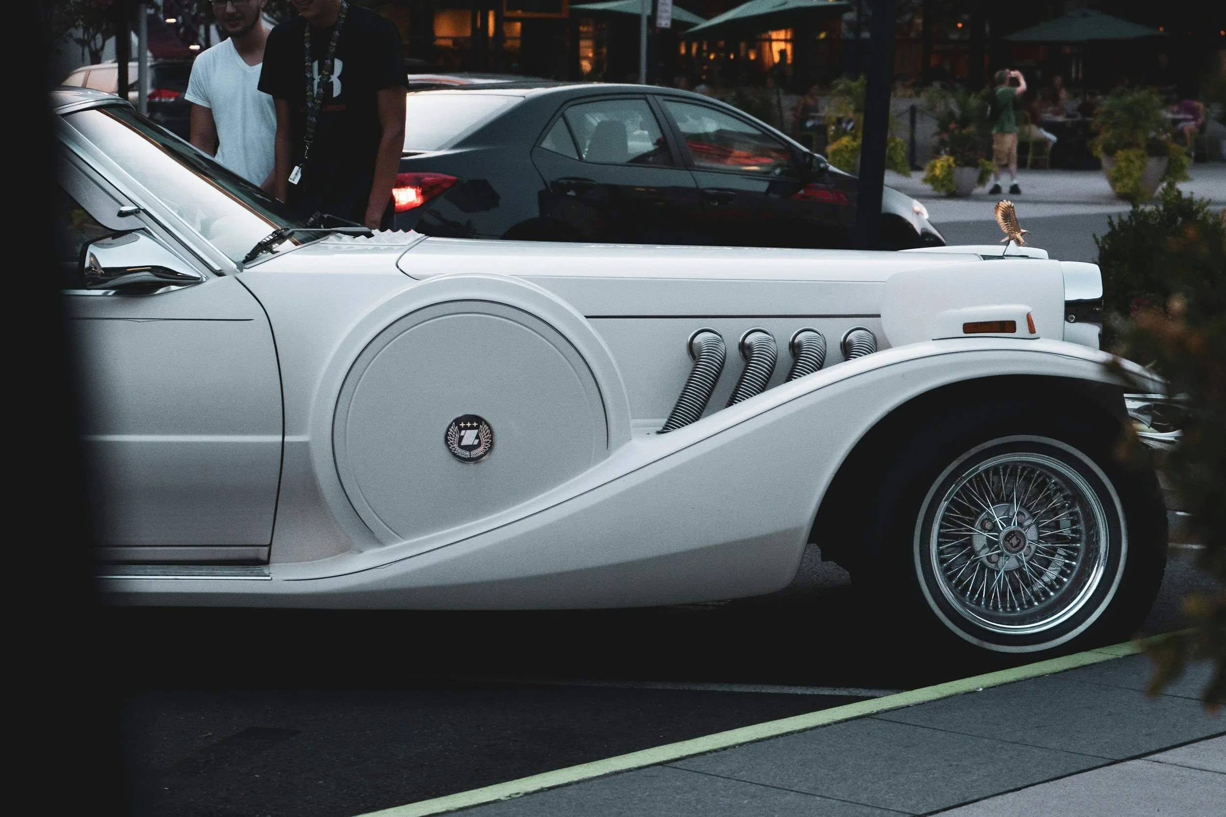 A white vintage car with a symbol on the door and exhaust pipes on the side, parked next to a black modern car in an outdoor setting during evening hours.