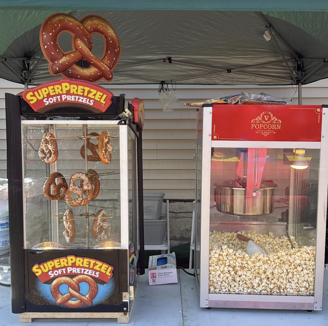 A popcorn machine and a soft pretzel vending machine at an outdoor event. The pretzel machine has several soft pretzels hanging inside, and a large pretzel decoration on top. The popcorn machine is filled with popcorn. There are containers and supplies behind the machines.