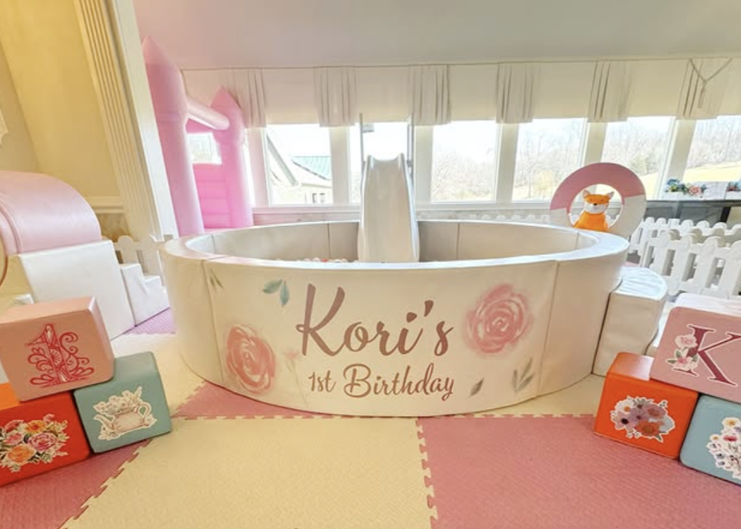 A decorated indoor space for Kori's 1st birthday with a white circular splash pad with a slide, surrounded by pink, blue, and orange blocks with floral and animal designs, and a window in the background.