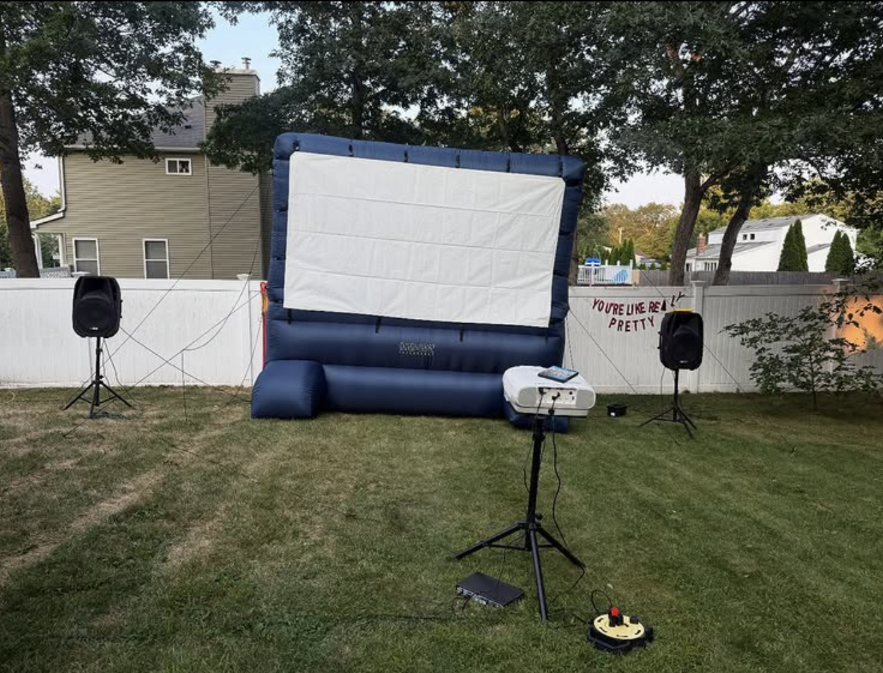 Outdoor backyard setup with a large inflatable movie screen, two speakers on stands, and a projector on a tripod on the grass. There is a small sign on the fence that reads 'You're like really pretty' and a white house behind trees.