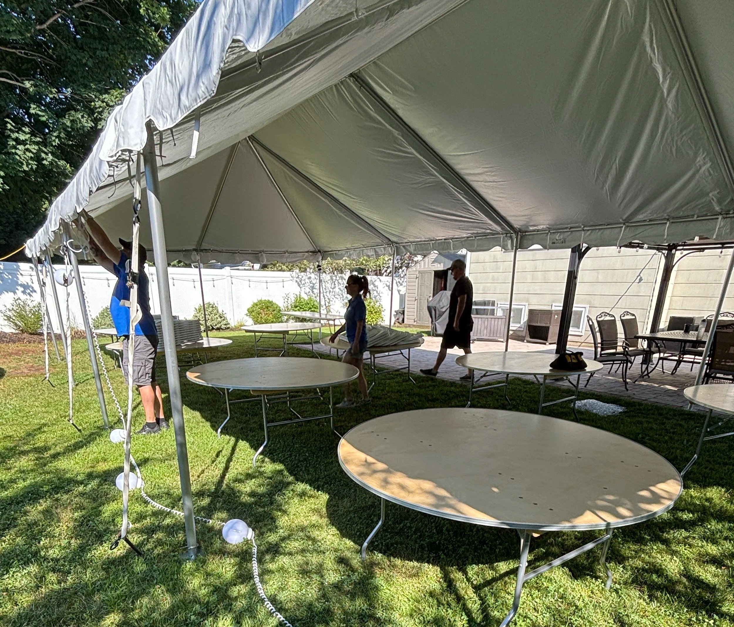 People setting up a white event tent outdoors with tables and chairs underneath, surrounded by greenery and a house in the background.