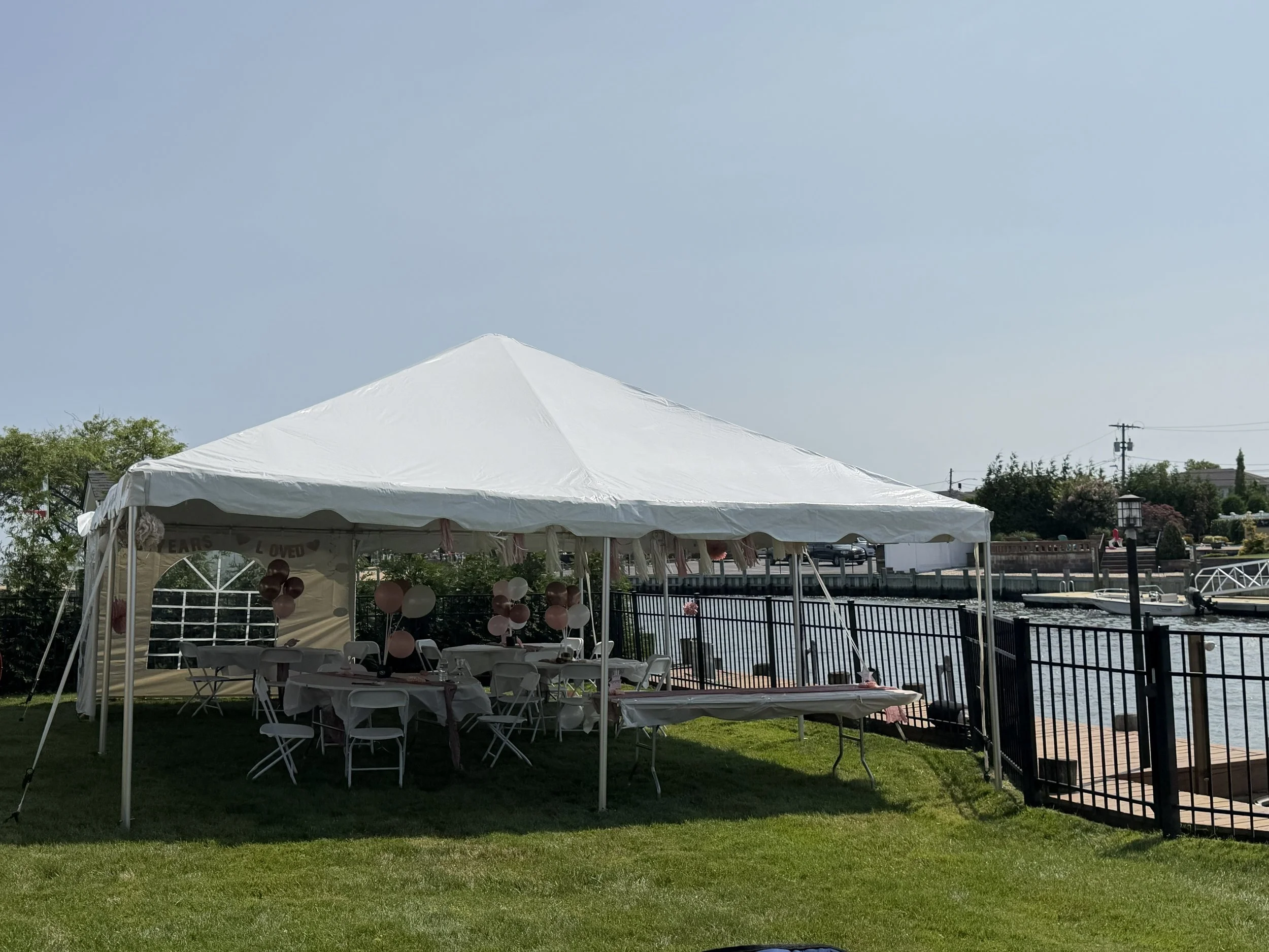 A white event tent set up outdoors near a body of water, with round balloons and chairs underneath, and a grassy area in the foreground.